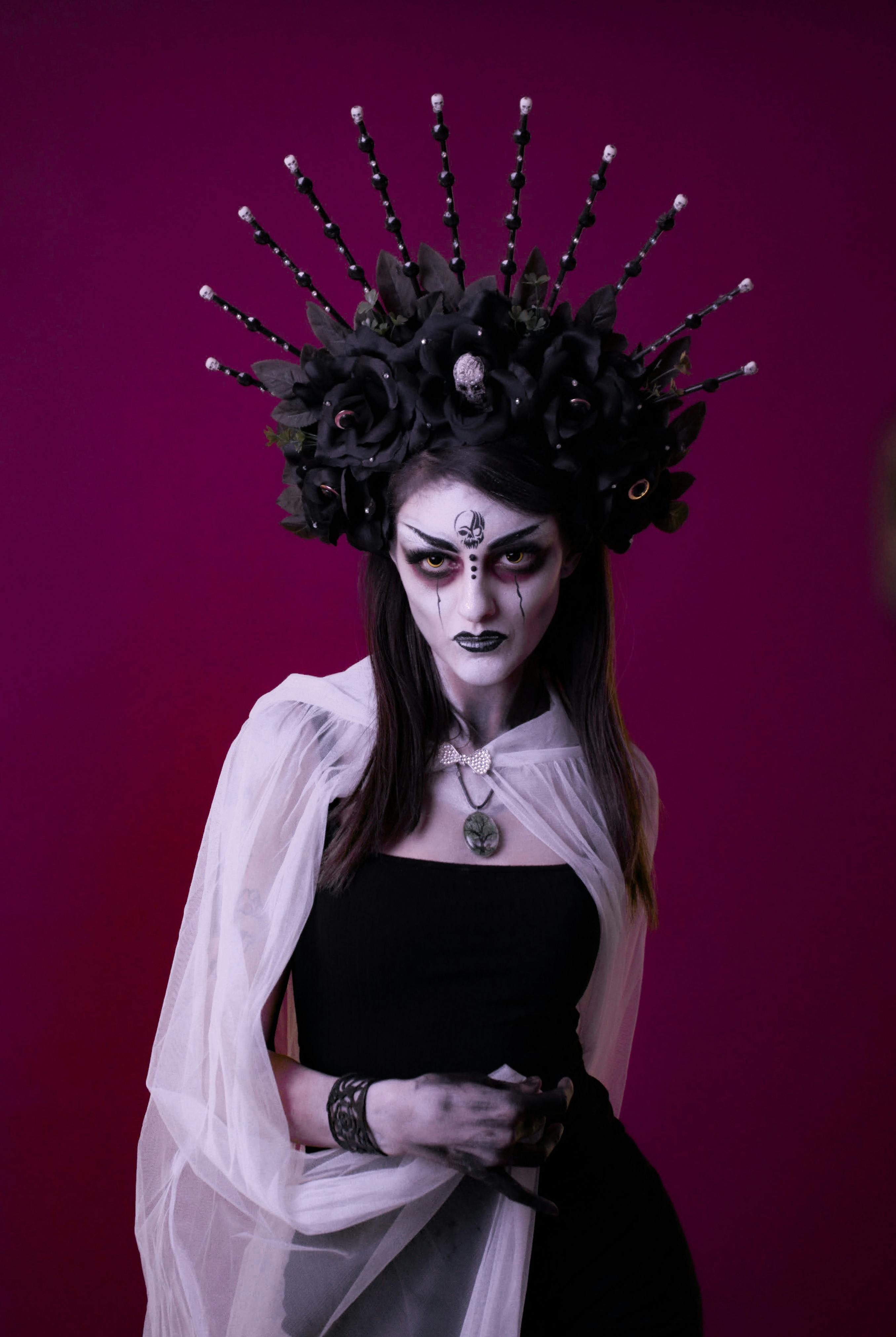 A woman in gothic makeup and attire with a floral headpiece against a purple backdrop.