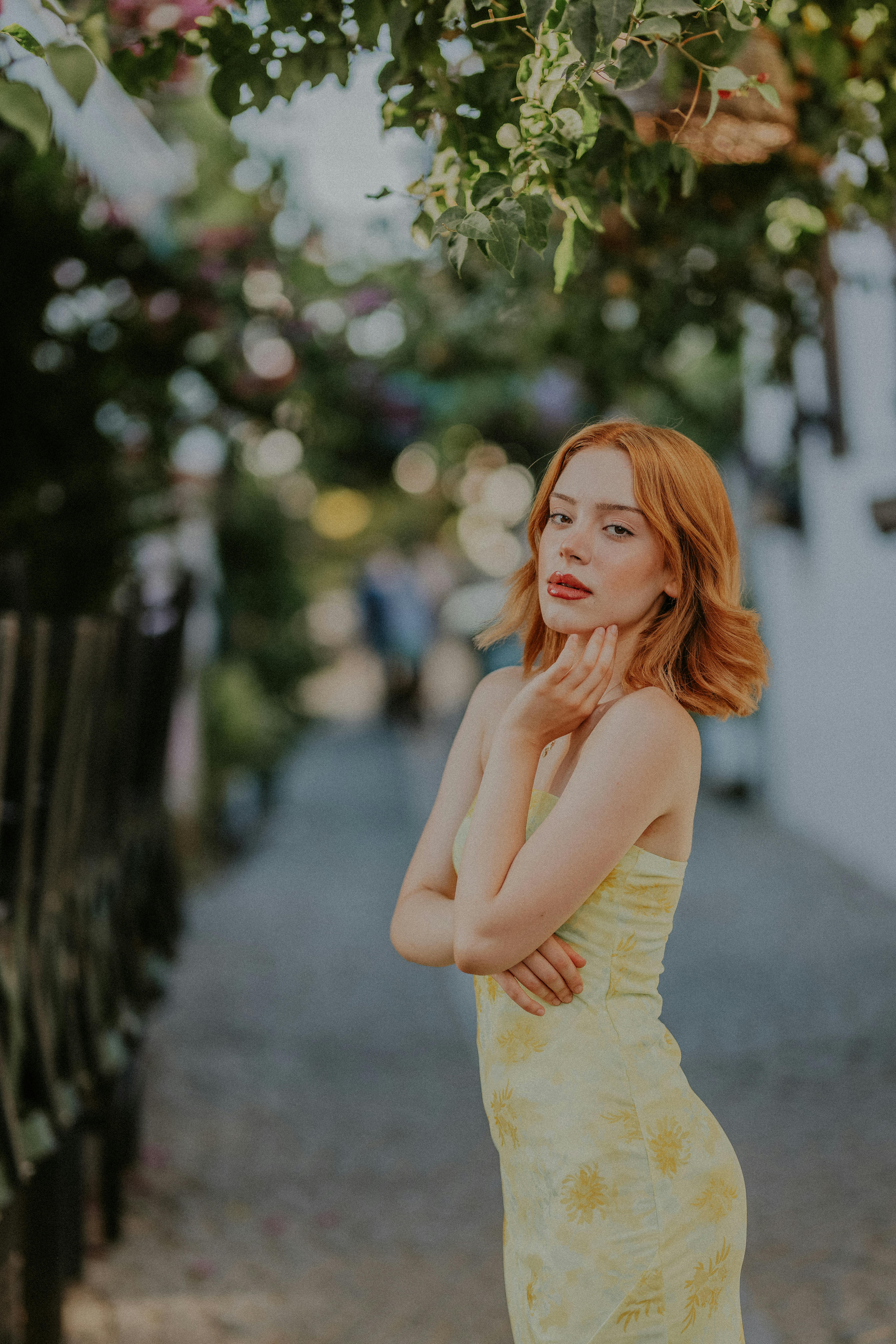 Red-haired woman in a yellow dress posing outdoors with floral background.