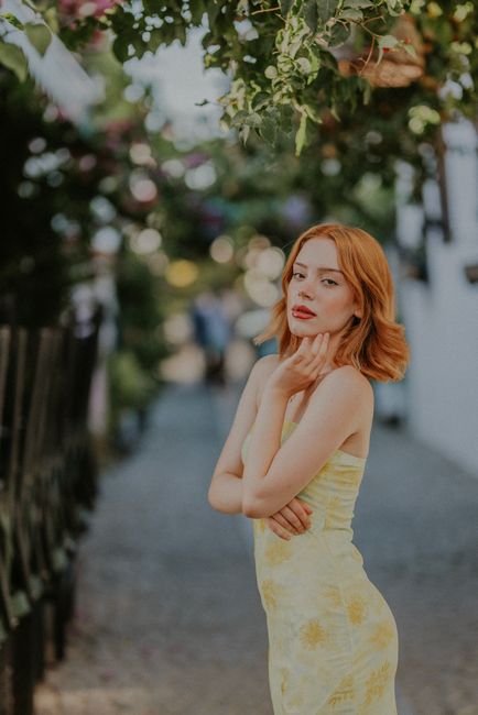 Red-haired woman in a yellow dress posing outdoors with floral background.