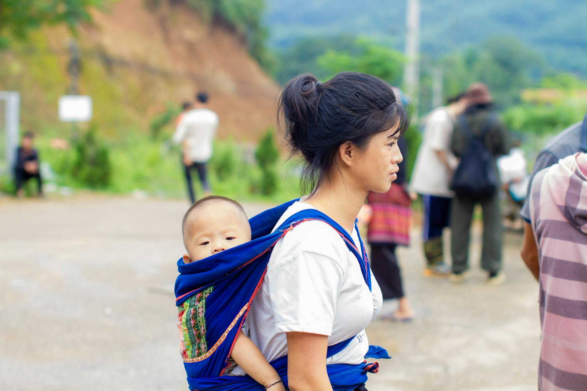 Young woman carrying baby on her back in a blue wrap outdoors, surrounded by nature.