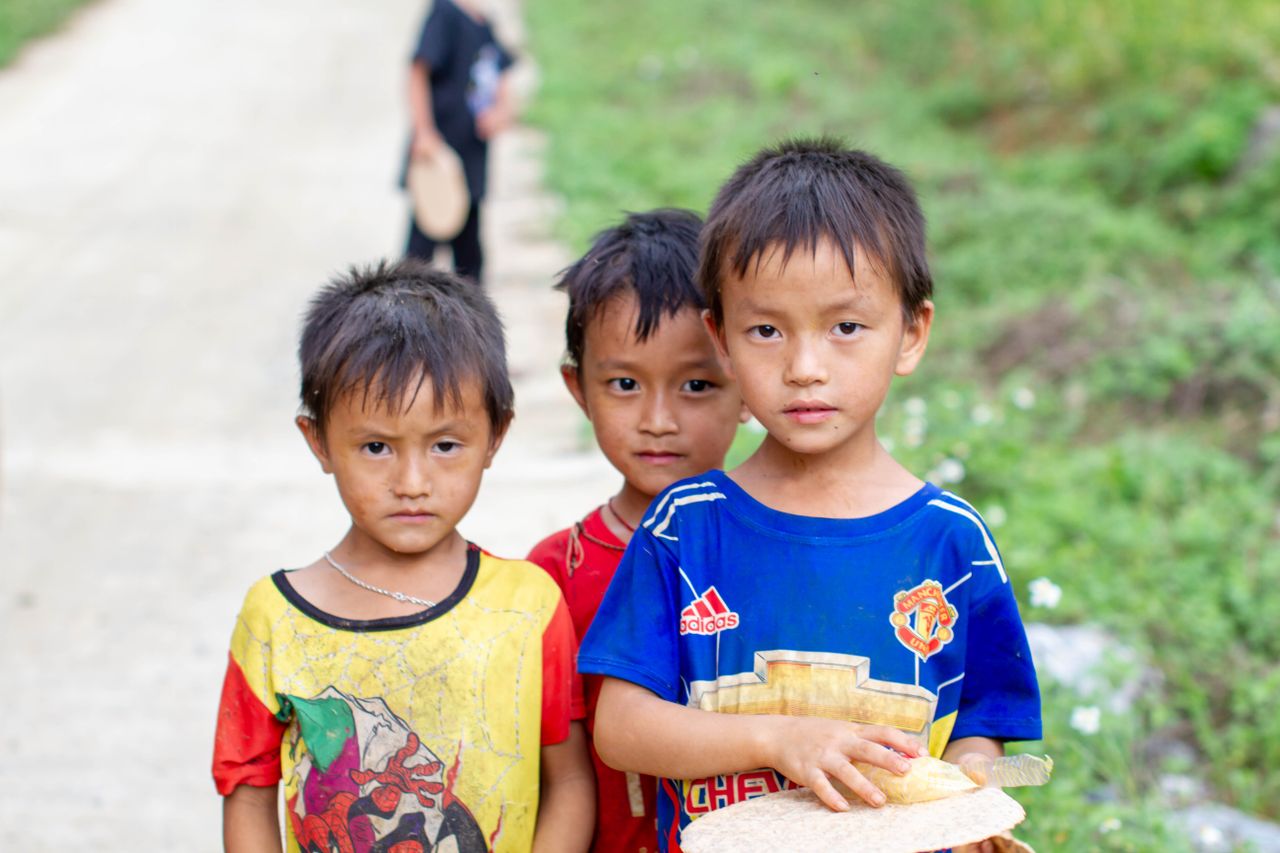 three young boys stand outdoors on a lush pathway looking directly at