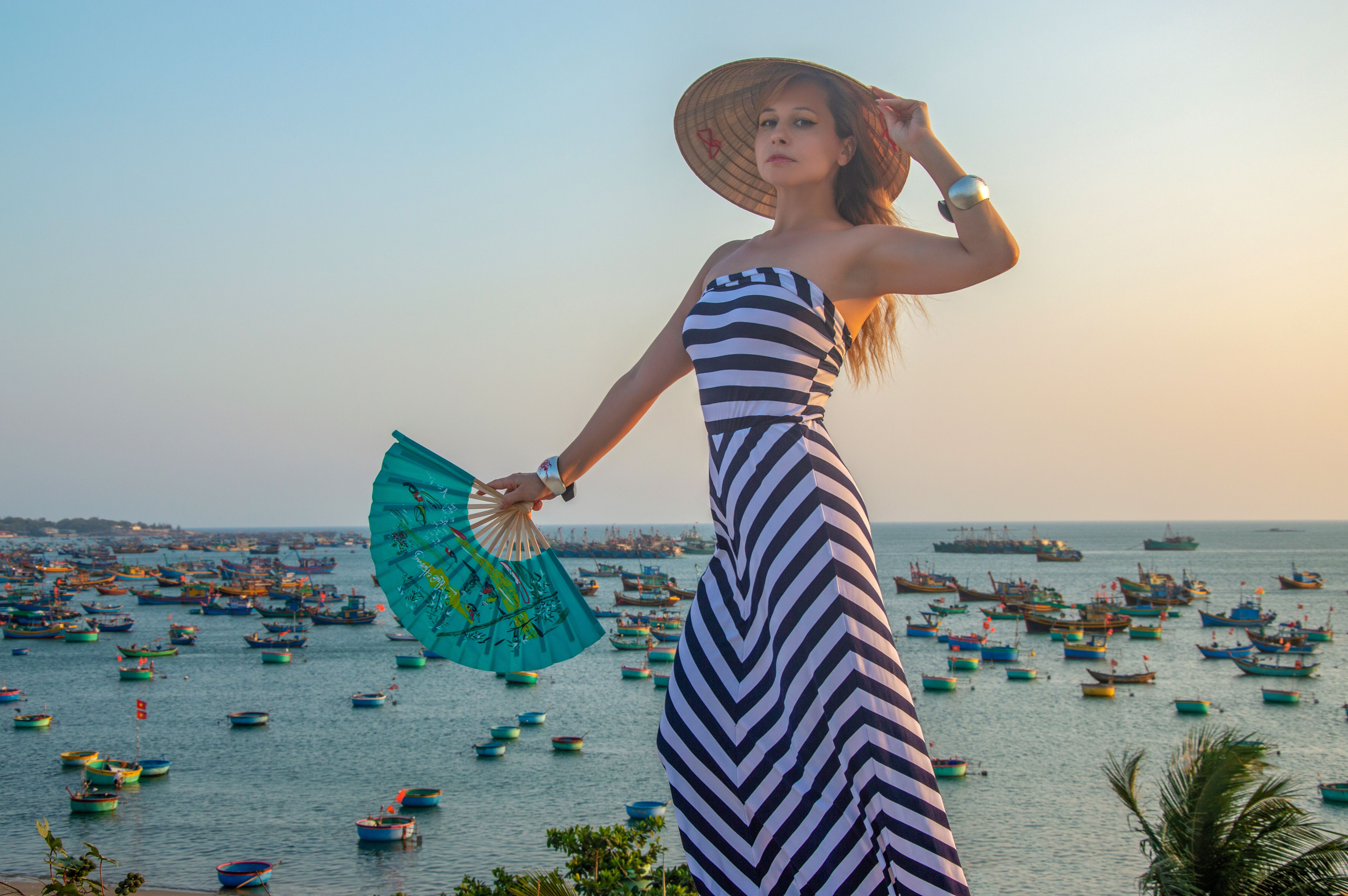 Mulher com chapéu tomando sol à beira mar, vestindo vestido listrado e segurando um leque colorido, em ambiente costeiro ao pôr do sol.
