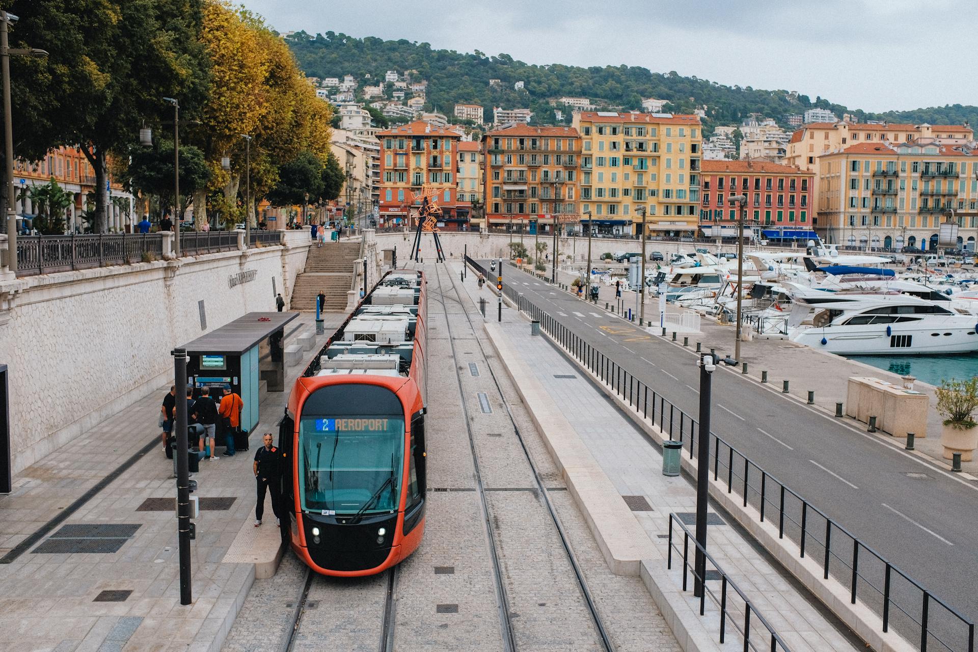 Red tram at a station in a vibrant coastal city with marina and colorful buildings.