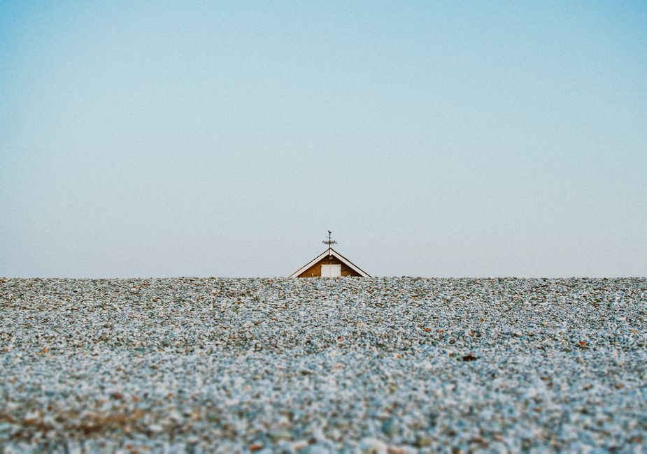 A single house roof peeks over a vast gravel field under a clear sky.