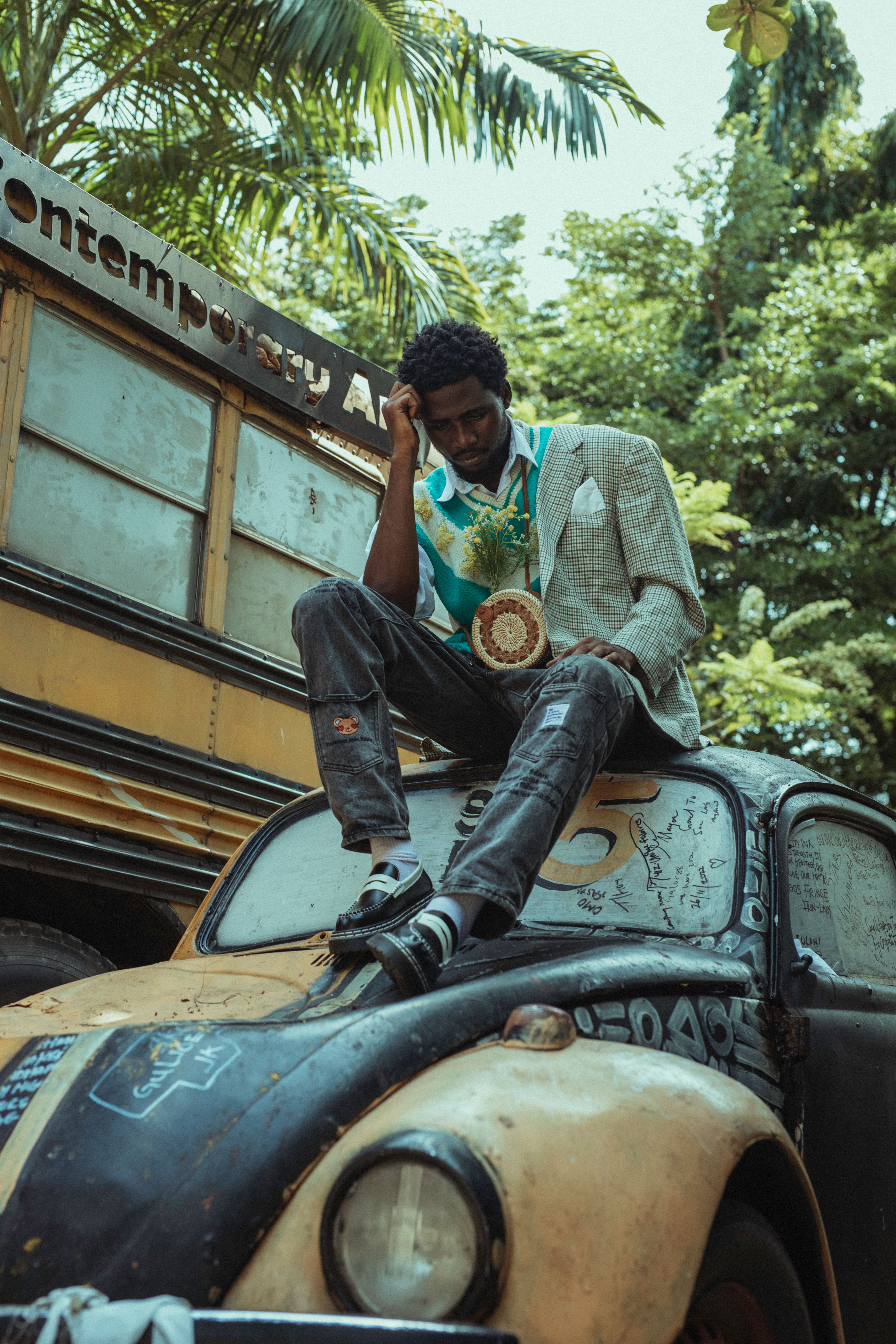Man in trendy outfit sits on old car near bus in lush outdoor setting.