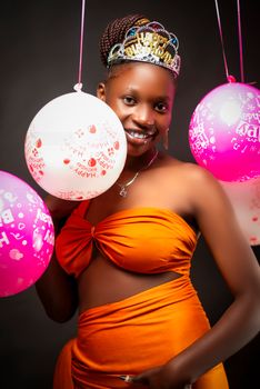 Woman in orange dress smiling with balloons and tiara at a party.