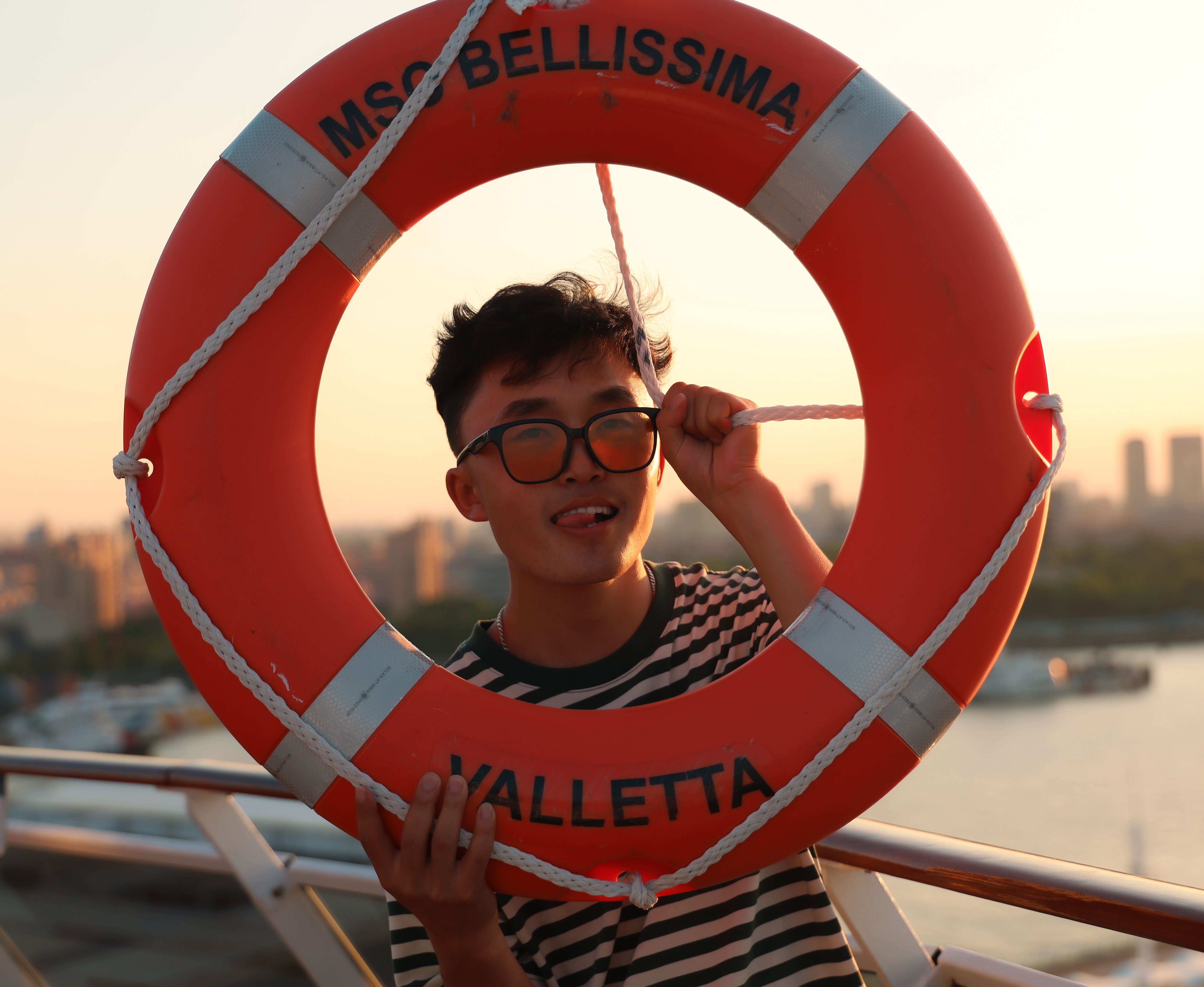 A young man playfully poses with a lifebuoy on a ship at sunset, featuring a vibrant and relaxed mood.