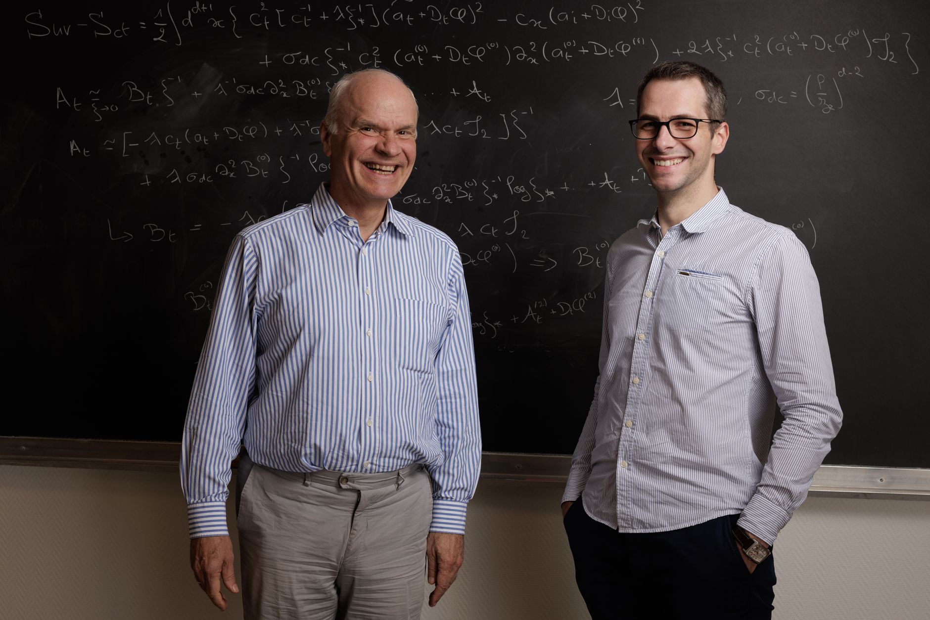 Two professors smiling in front of a blackboard filled with complex mathematical equations.