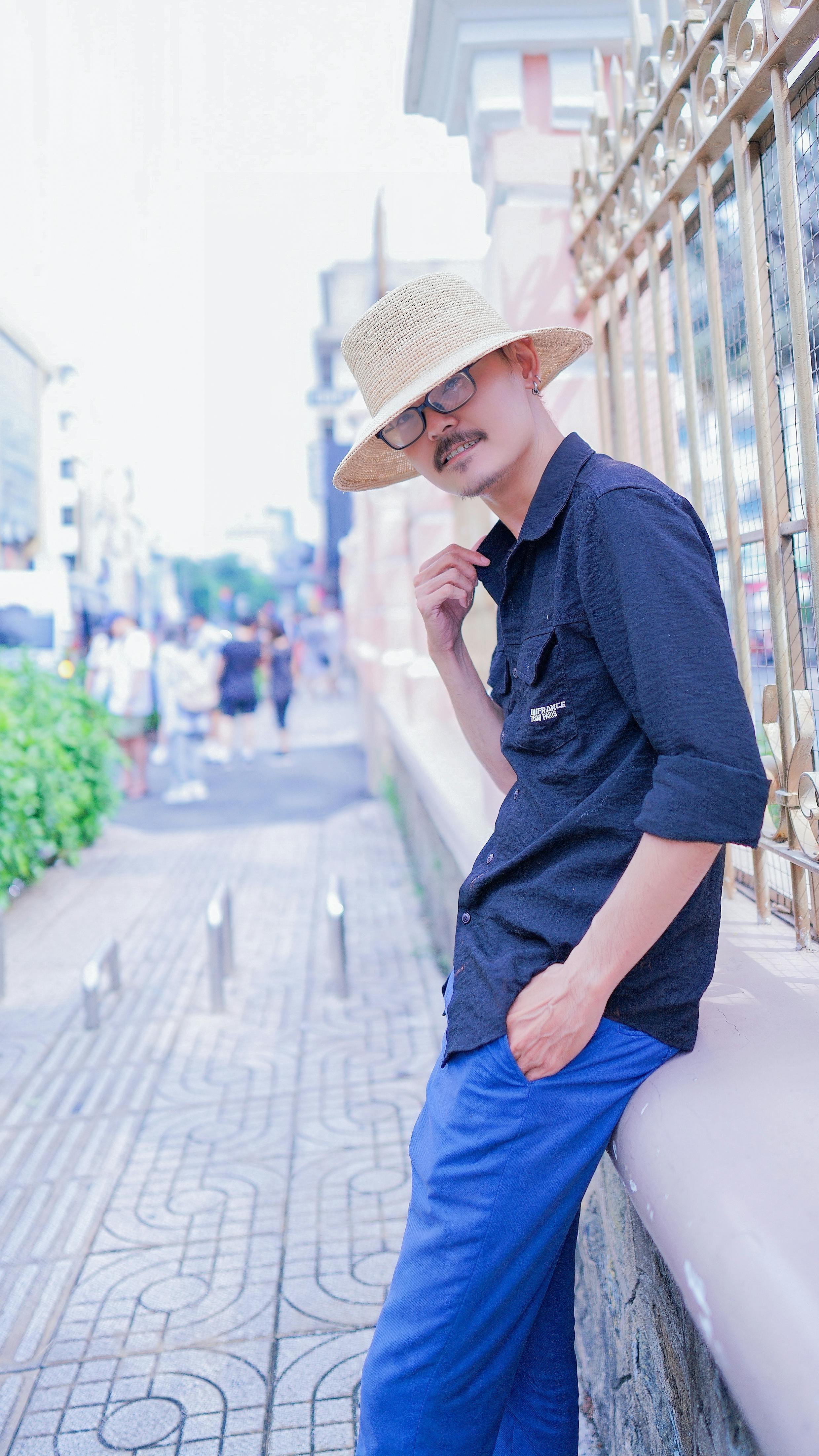 A fashionable adult man wearing a straw hat and sunglasses, posing outdoors by a fence.