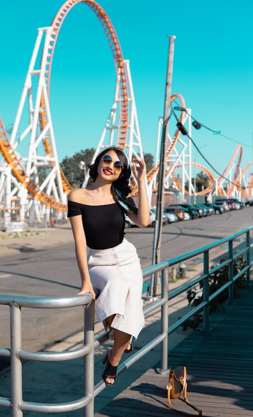 Woman in Black Off-shoulder Top, White Skirt, and Black High-heeled Mules Sitting on Silver Balustrade With Roller Coaster Background