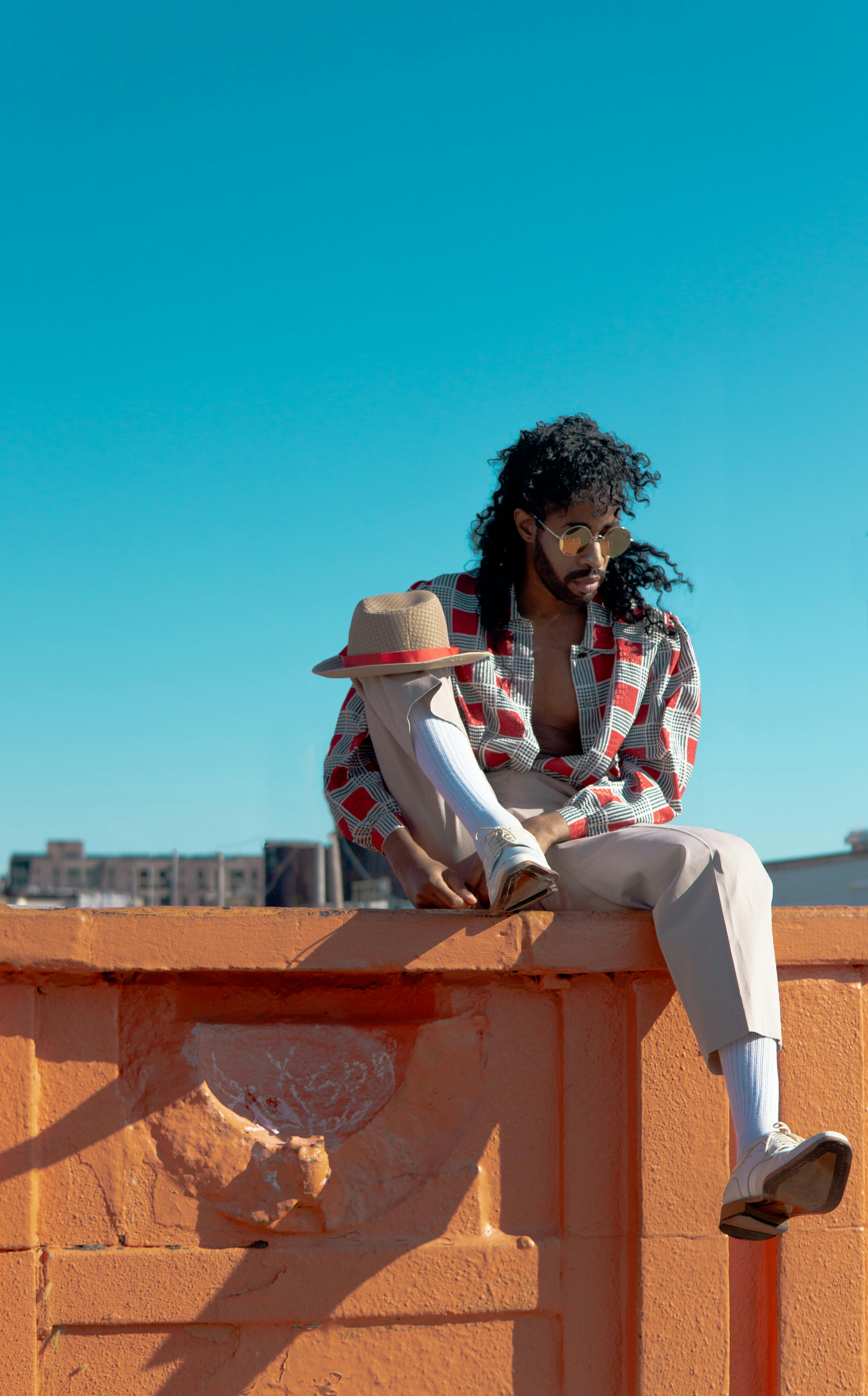 Fashionable man with curly hair posing on an orange wall under blue skies, embodying summer style.