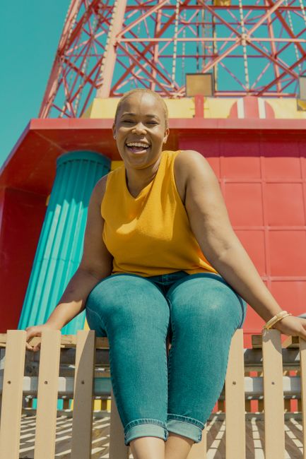 A cheerful woman in casual attire smiles outdoors with a colorful structure in the background.