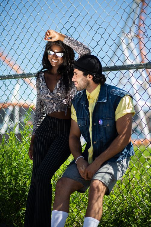 Free Fashionable couple posing by a chain-link fence on a sunny day, showcasing casual street style. Stock Photo