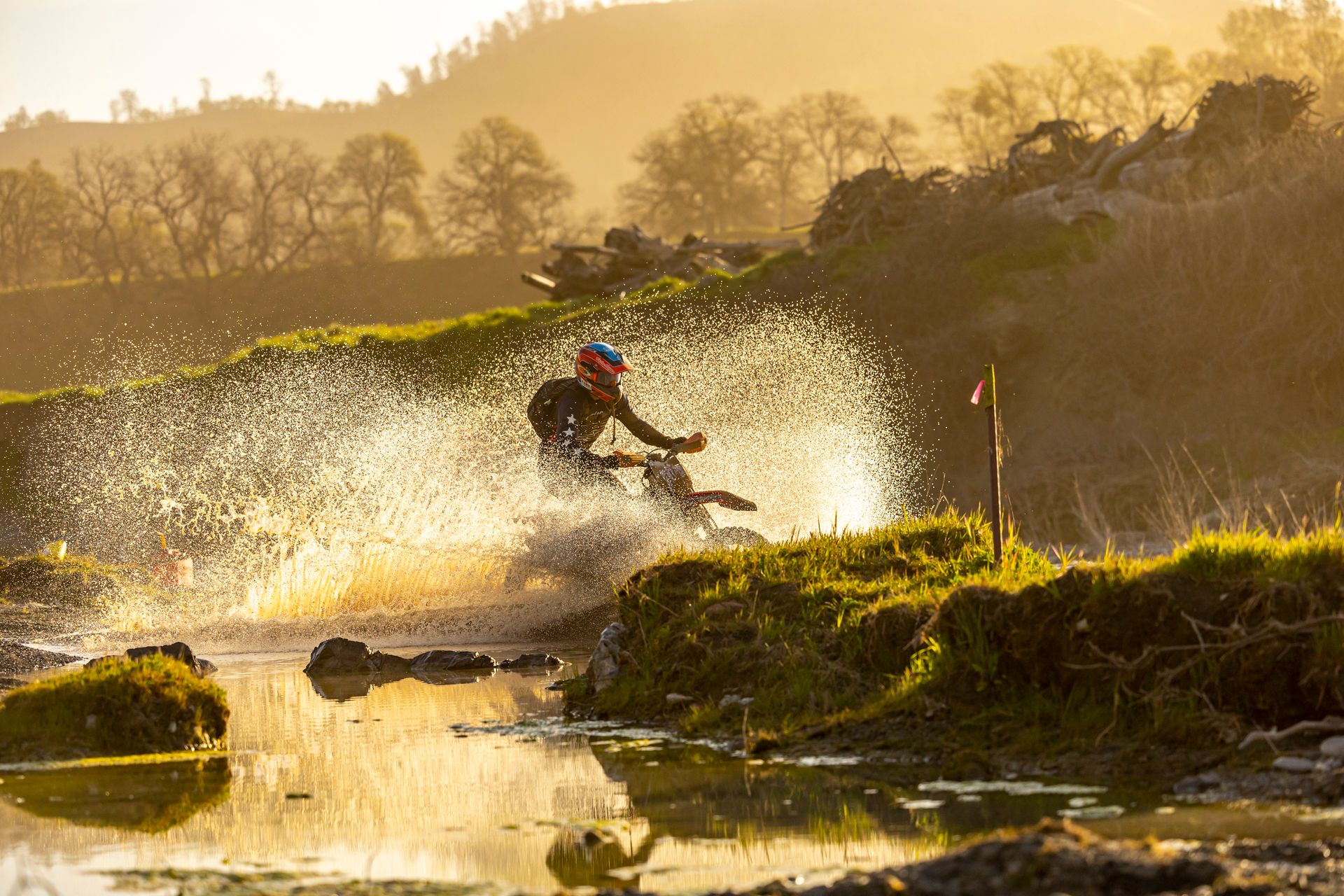 Dramatic shot of a motocross rider splashing through water with a scenic sunset backdrop.