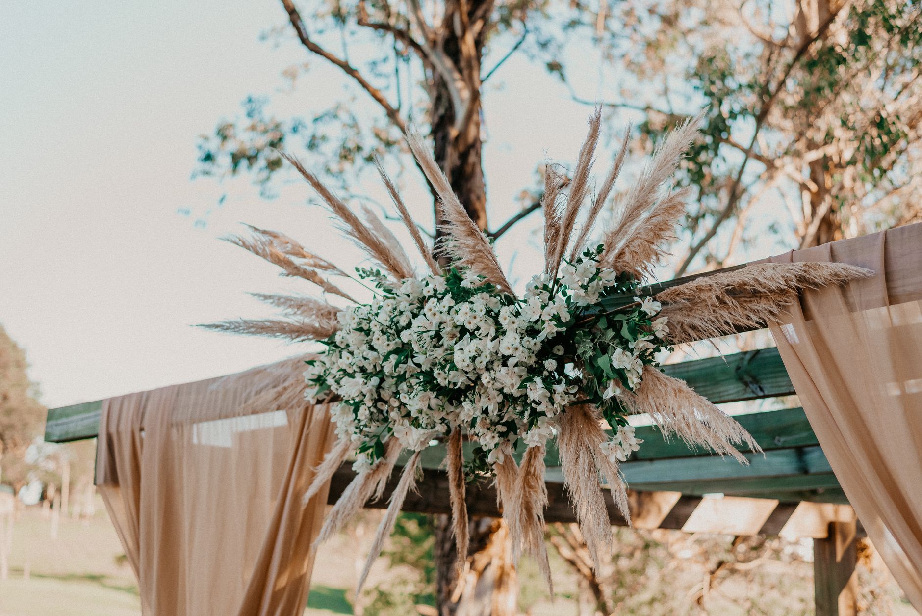 A beautifully decorated outdoor wedding arbor with flowers and pampas grass under a sunny sky.