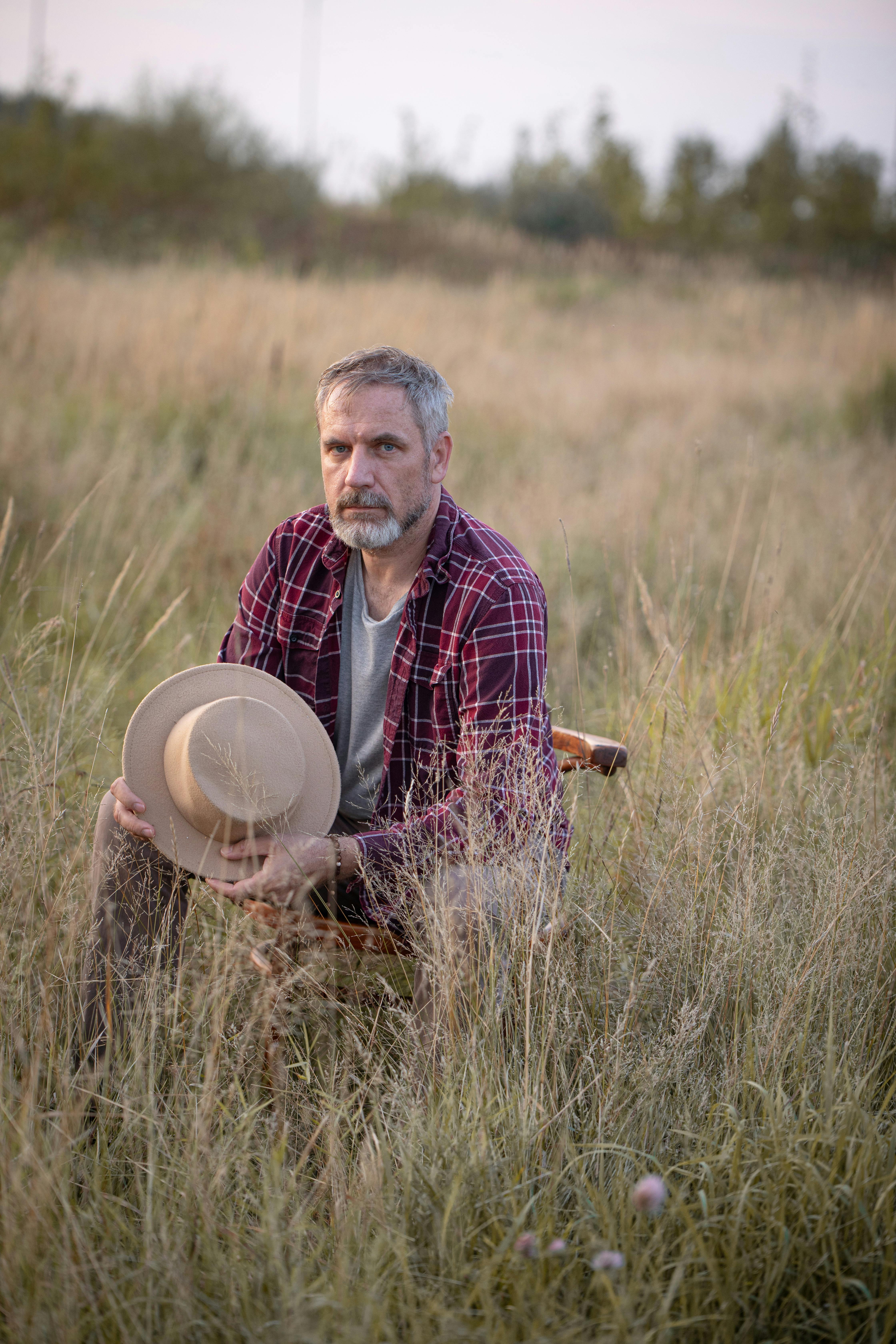 Elderly man holding hat sitting in serene grassy field during sunset, showcasing peace and contemplation.