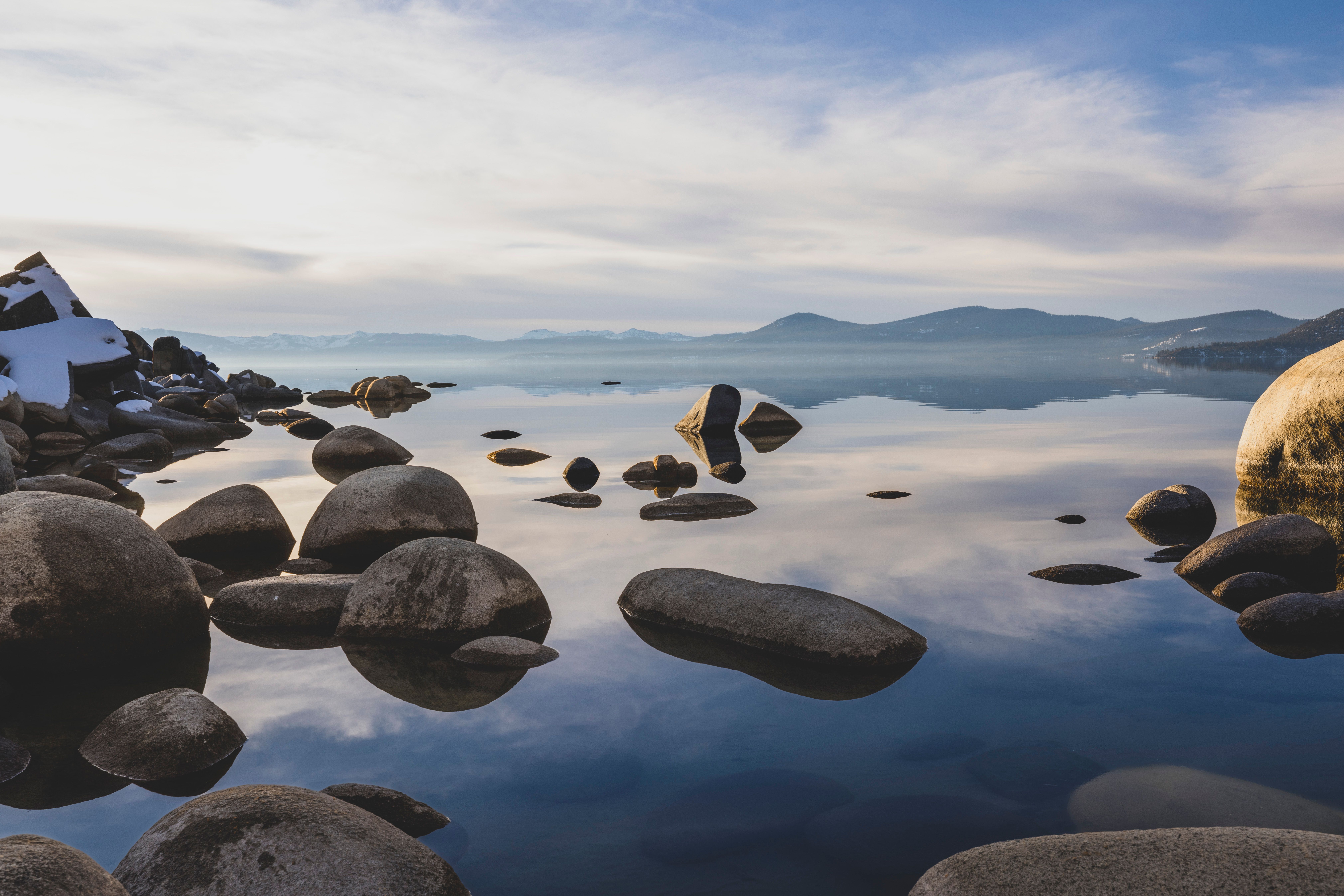 Tranquil lake scene with smooth stones and a clear sky reflecting on the water's surface.