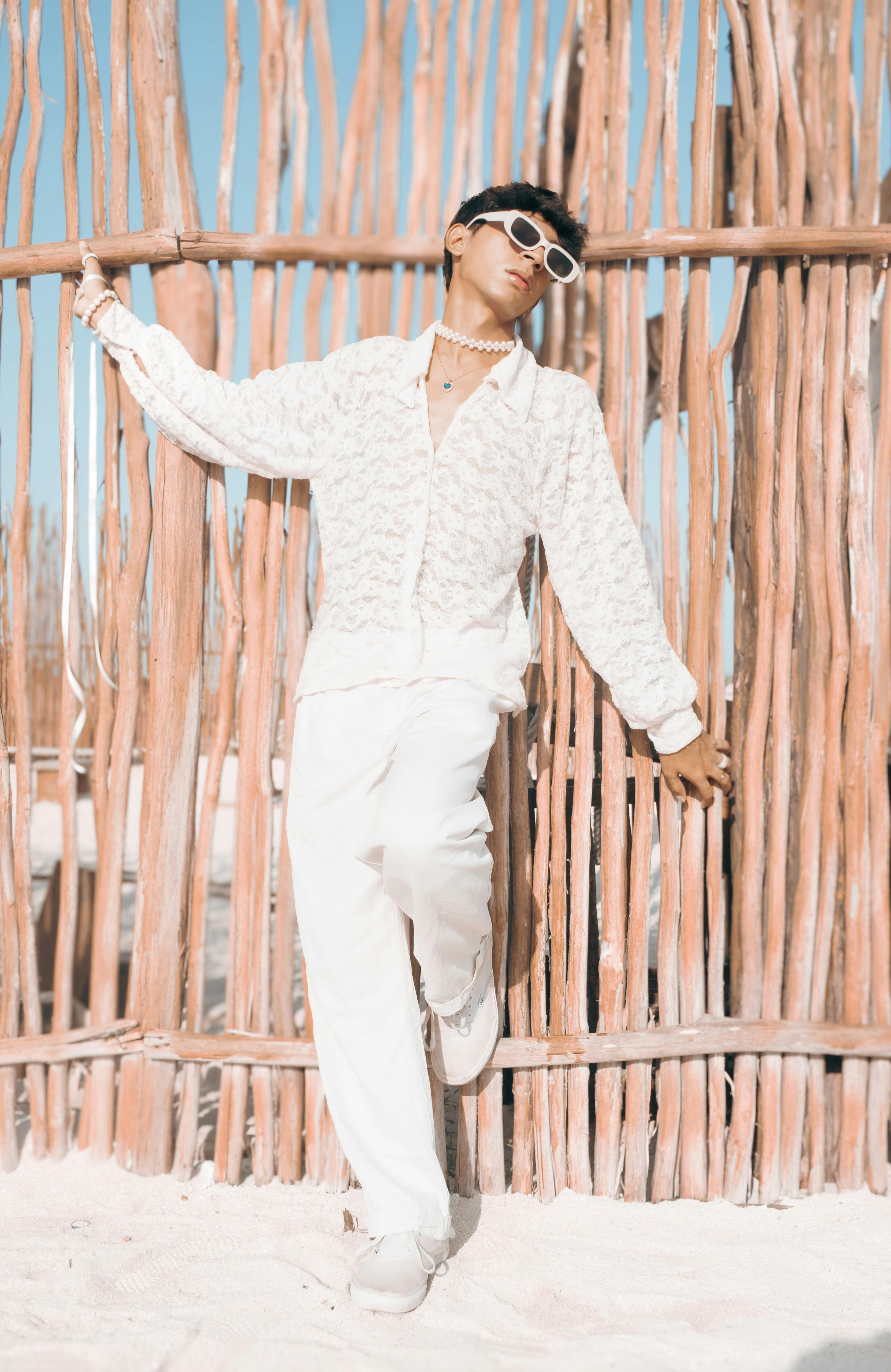 Fashionable young man in white attire leaning against rustic wooden fence under blue sky.