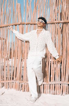 Fashionable adult posing on beach with sunglasses and in stylish white attire, set against blue sky.