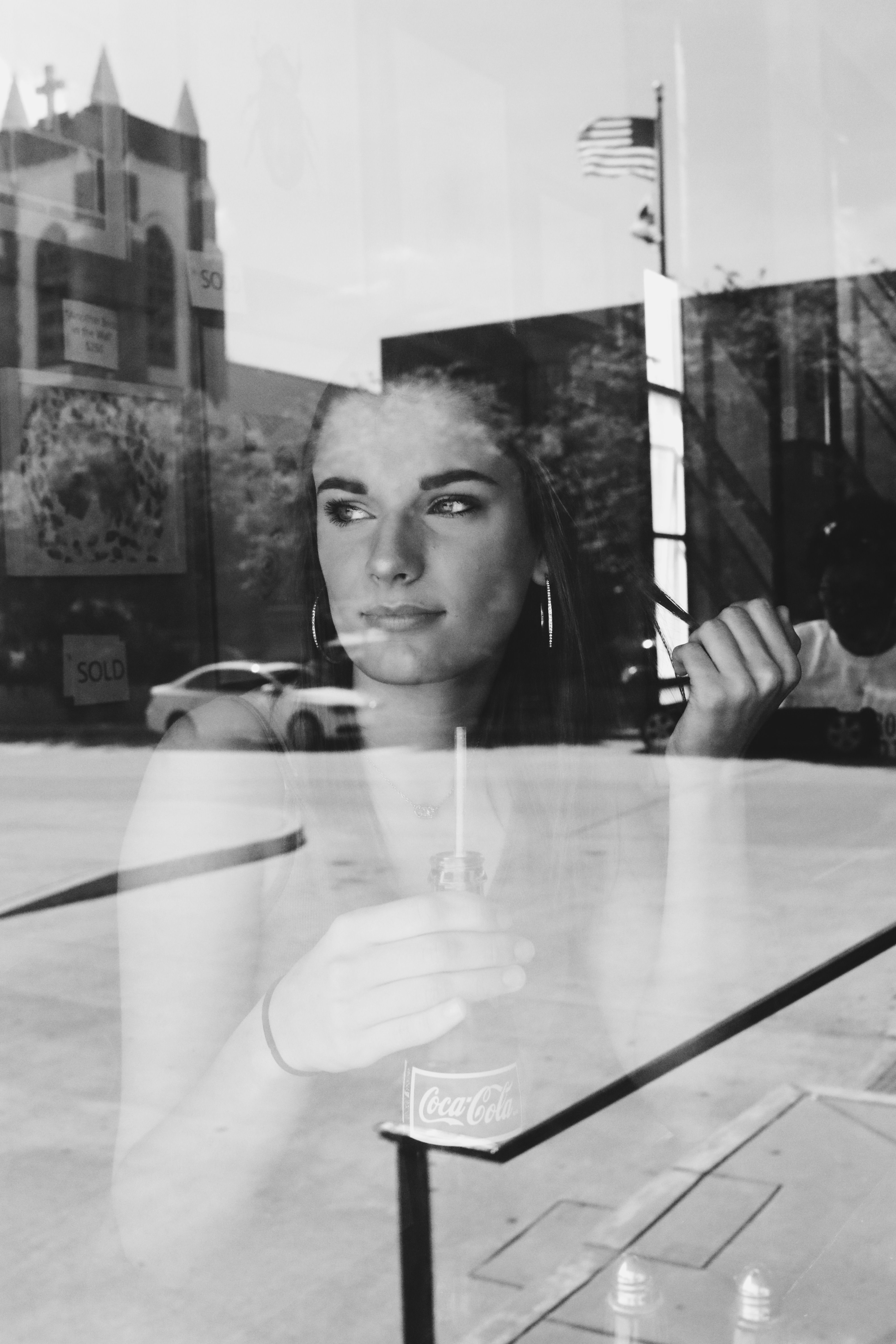 Black and white photo of a woman contemplating through a cafe window, capturing reflections and city life.