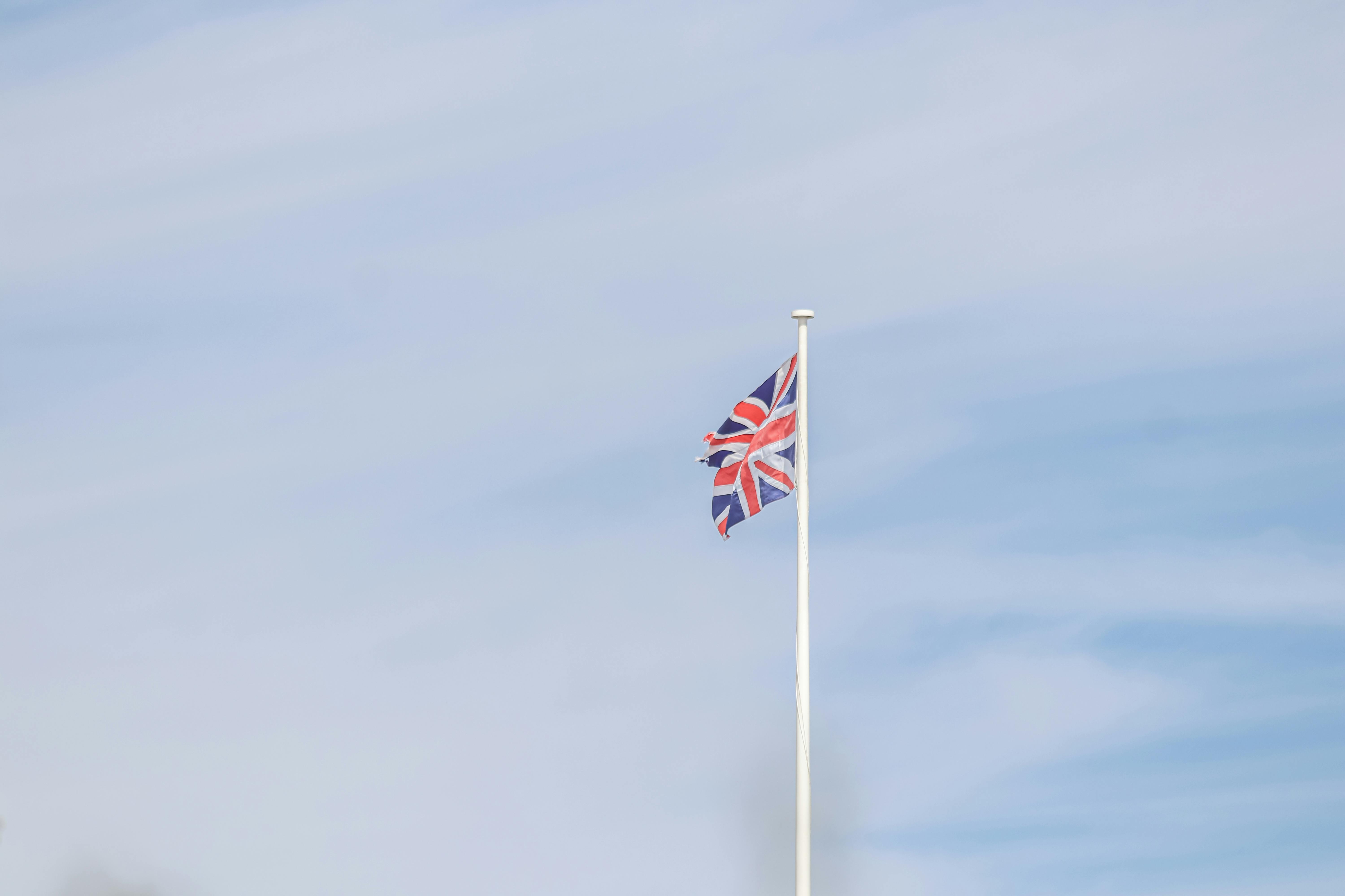 Union Jack flag on a pole against a light blue cloudy sky, symbolizing UK pride.