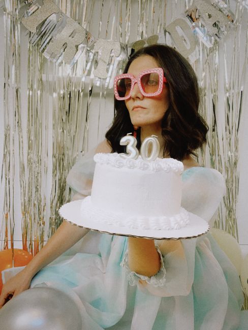 Woman wearing pink glasses holding a 30th birthday cake at a party.