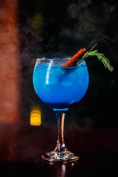 A vibrant blue cocktail garnished with cinnamon and rosemary in a dim setting.