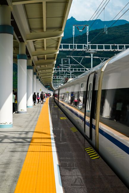 A modern high-speed train awaits at a bustling railway station with a mountain backdrop.