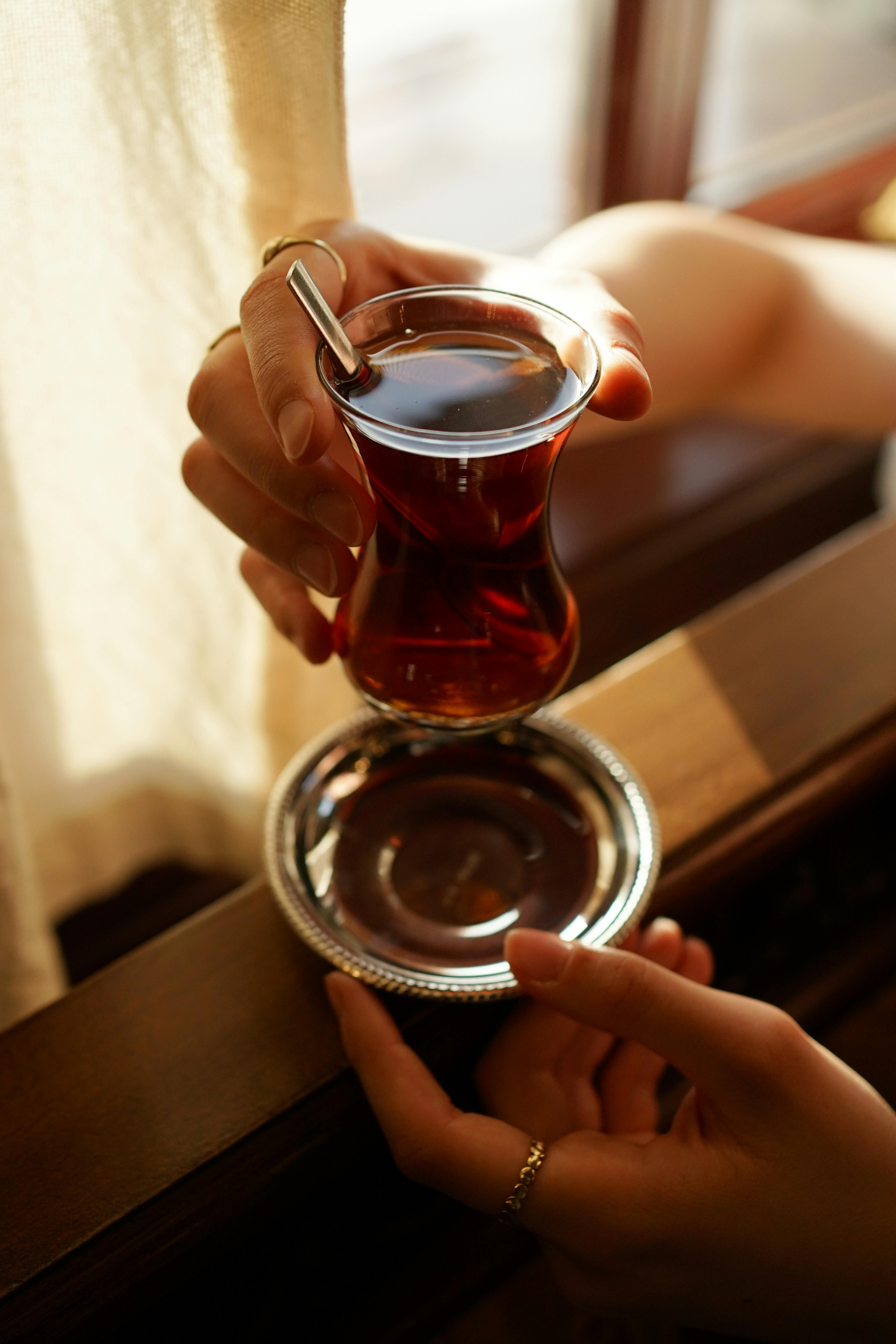 A woman's hands holding a glass of Turkish tea with a saucer, bathed in warm sunlight by a window.