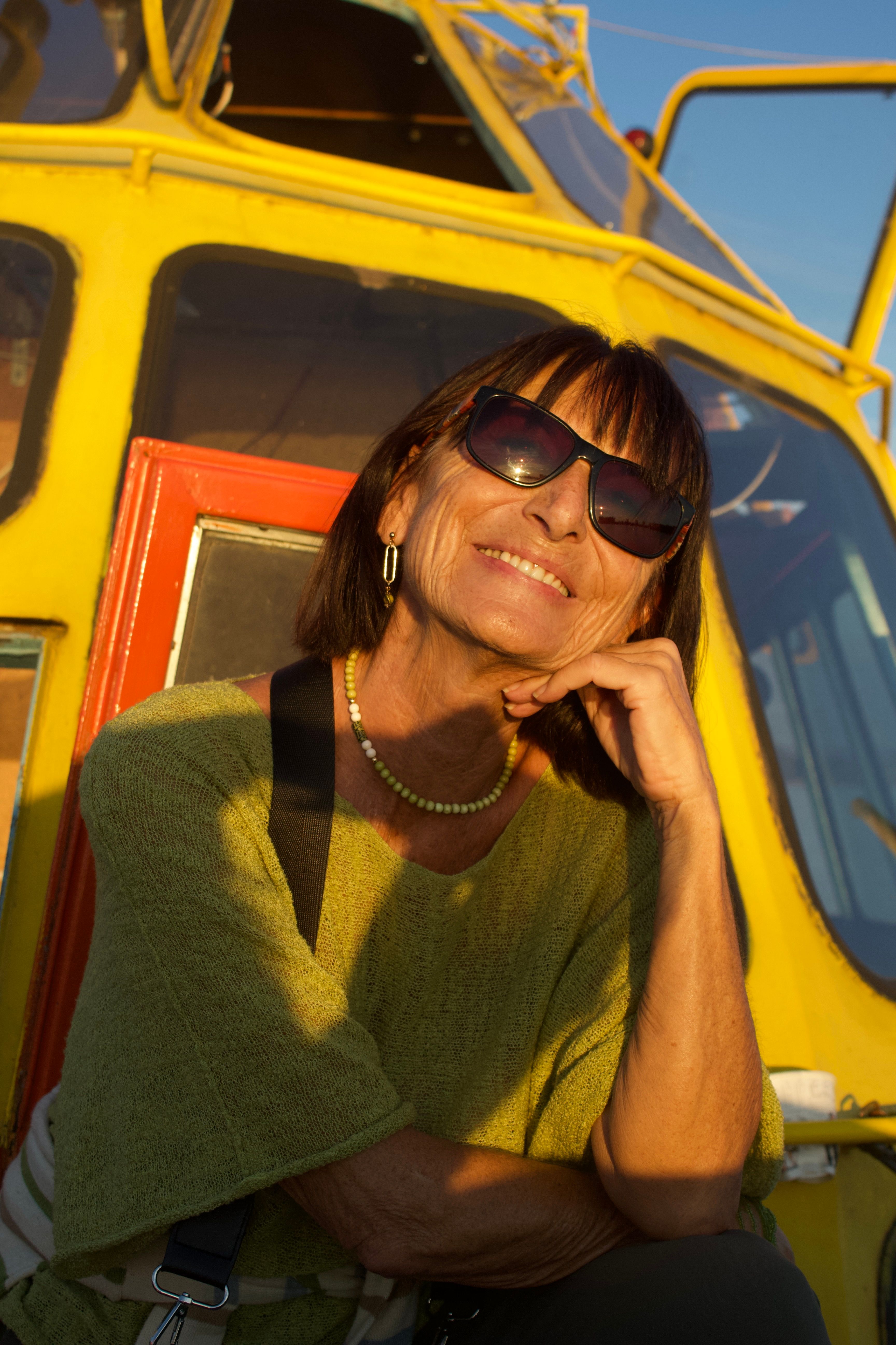 Smiling woman in sunglasses on a boat deck during sunset, wearing a green sweater.