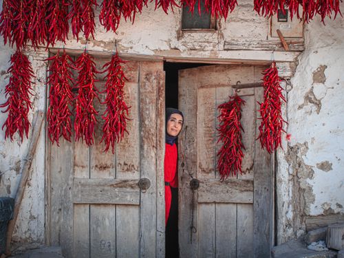 Free Woman in traditional attire peeks through rustic doorway adorned with hanging red peppers. Stock Photo