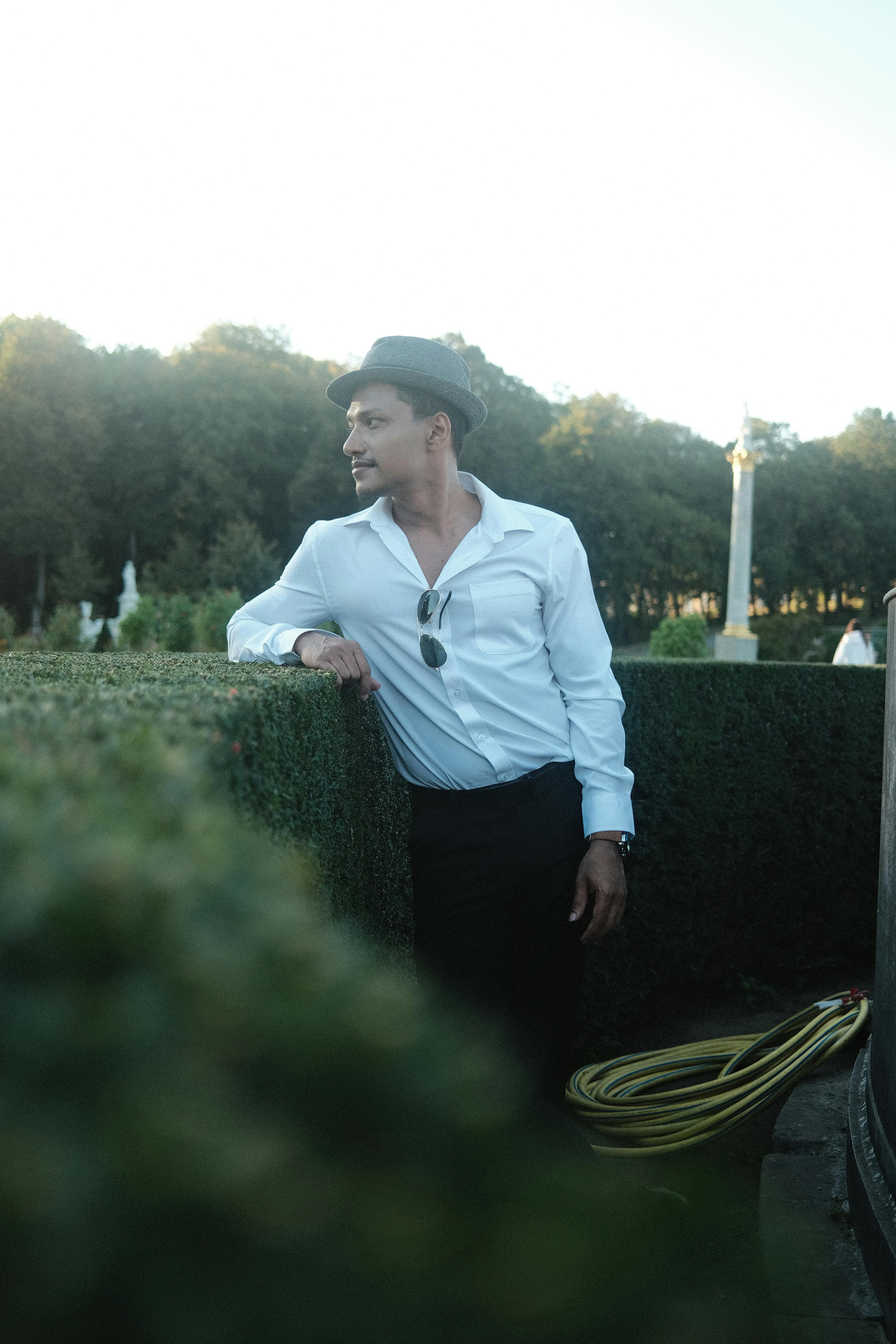 A man in a white shirt and hat leaning against a hedge in a garden.