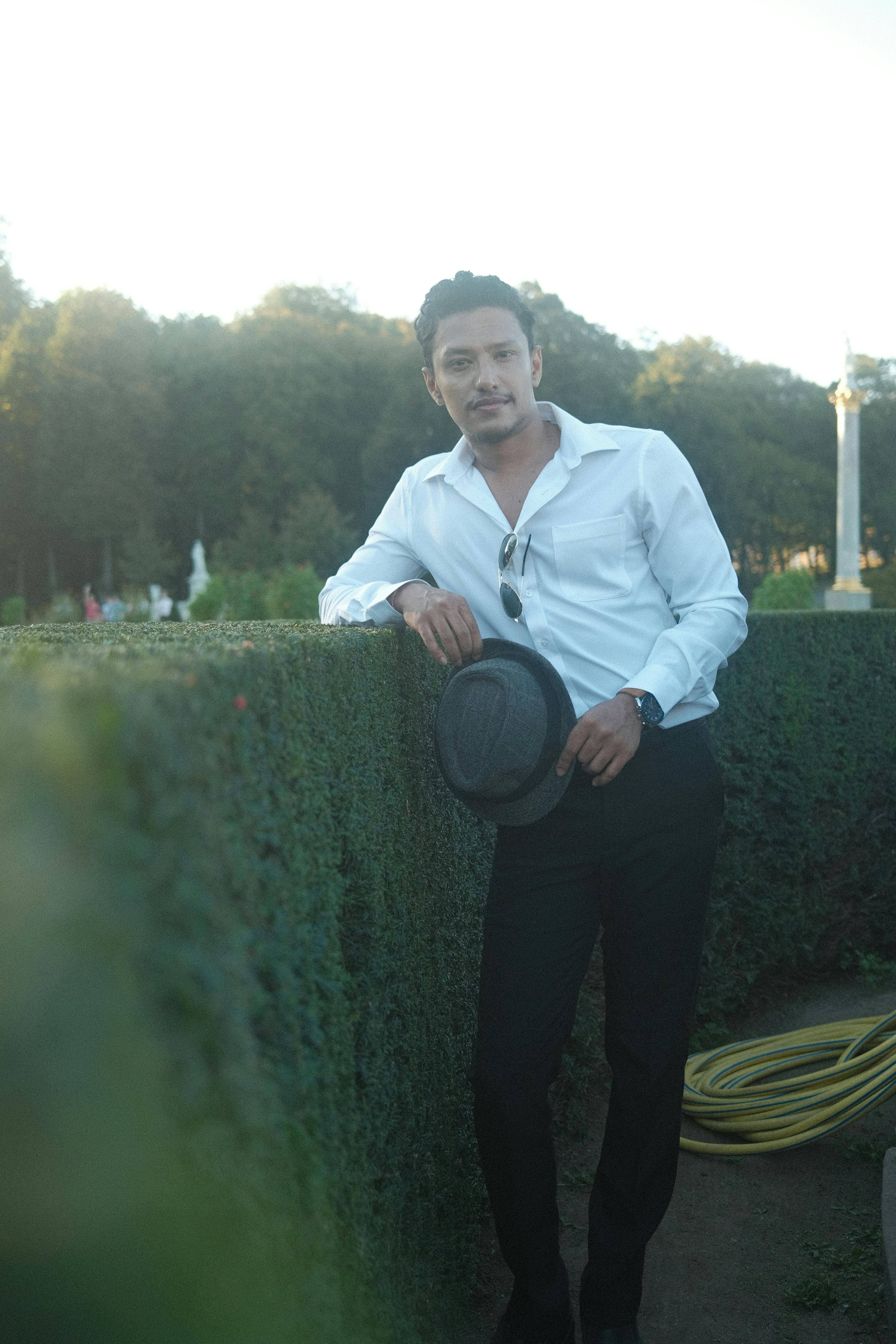 Man leaning on hedge in an urban garden setting, exuding style and elegance at dusk.