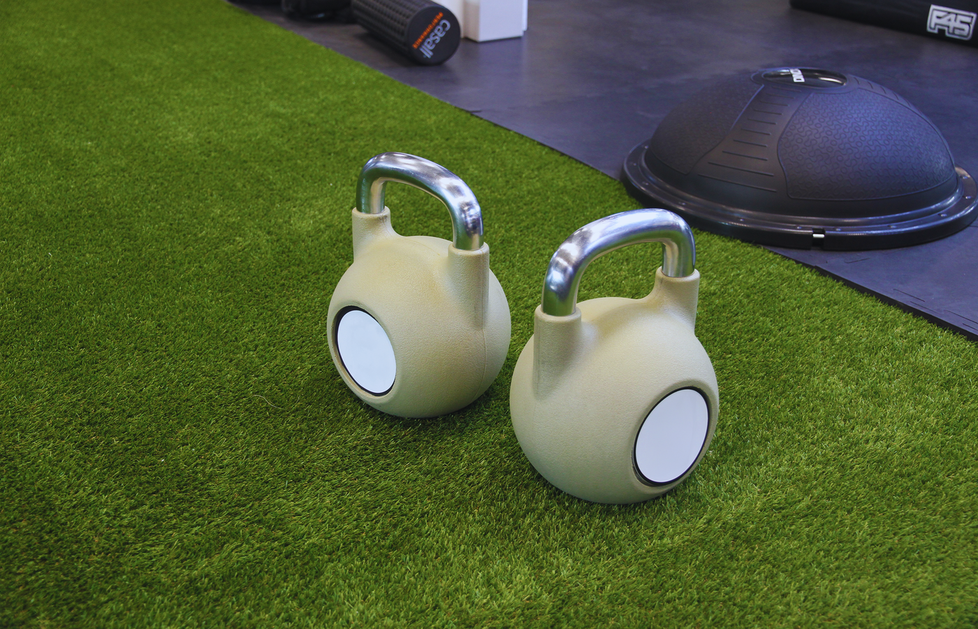Two kettlebells on a green artificial grass floor in an indoor gym, ideal for strength training.