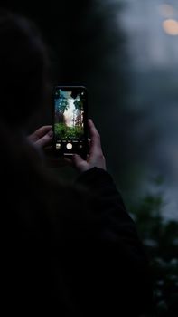 Person taking photo of trees with smartphone at twilight, capturing serene nature view.