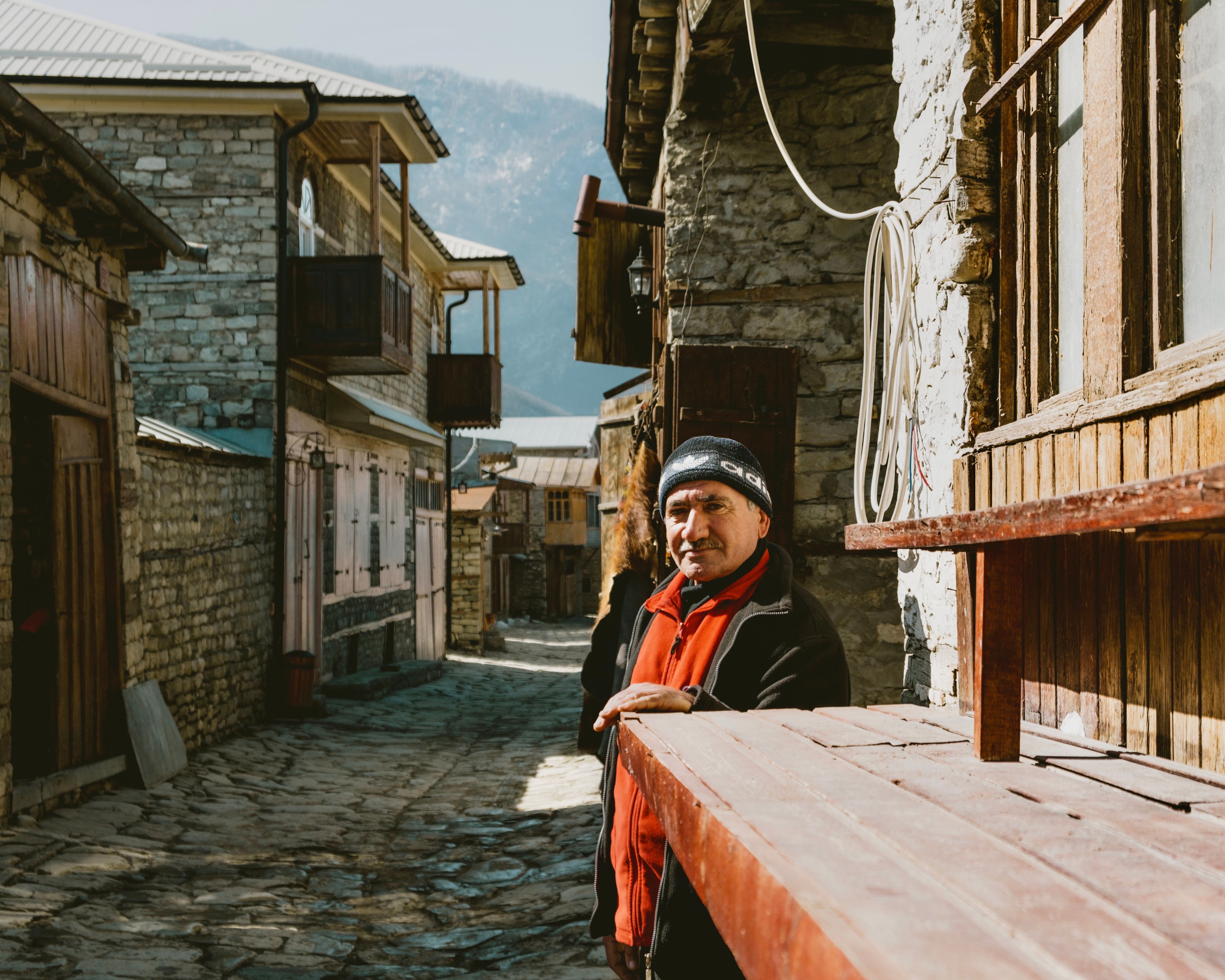 Elderly man in warm attire stands in a historic stone village street on a bright day.