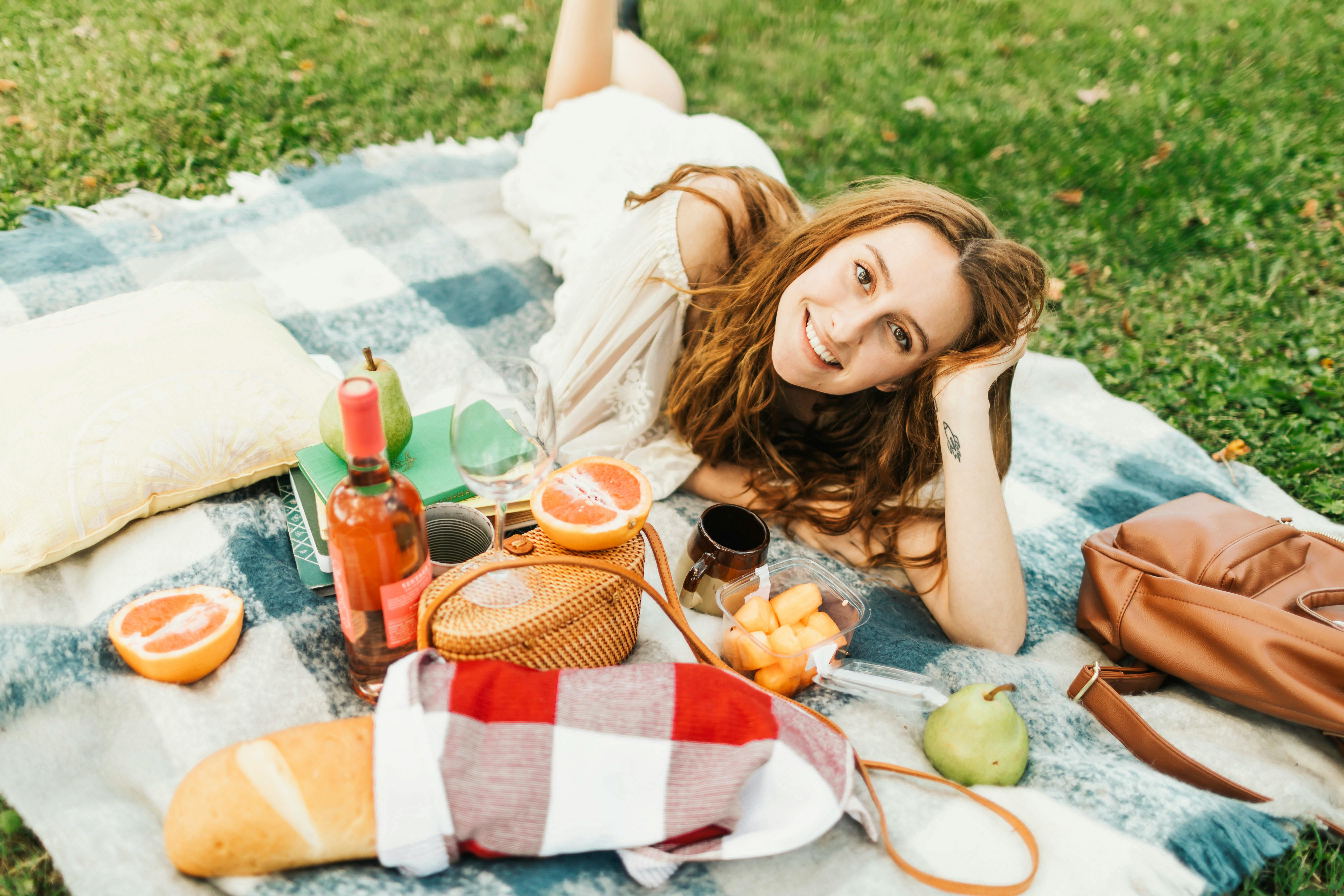 一位女士在阳光明媚的公园里野餐，她吃着水果，喝着葡萄酒，对着镜头微笑