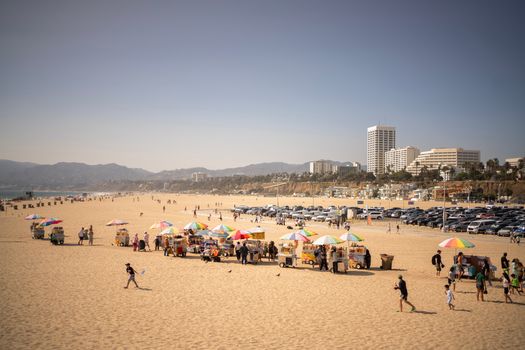 Sunny day at Santa Monica Beach with colorful vendor stalls and people strolling the sandy shore.