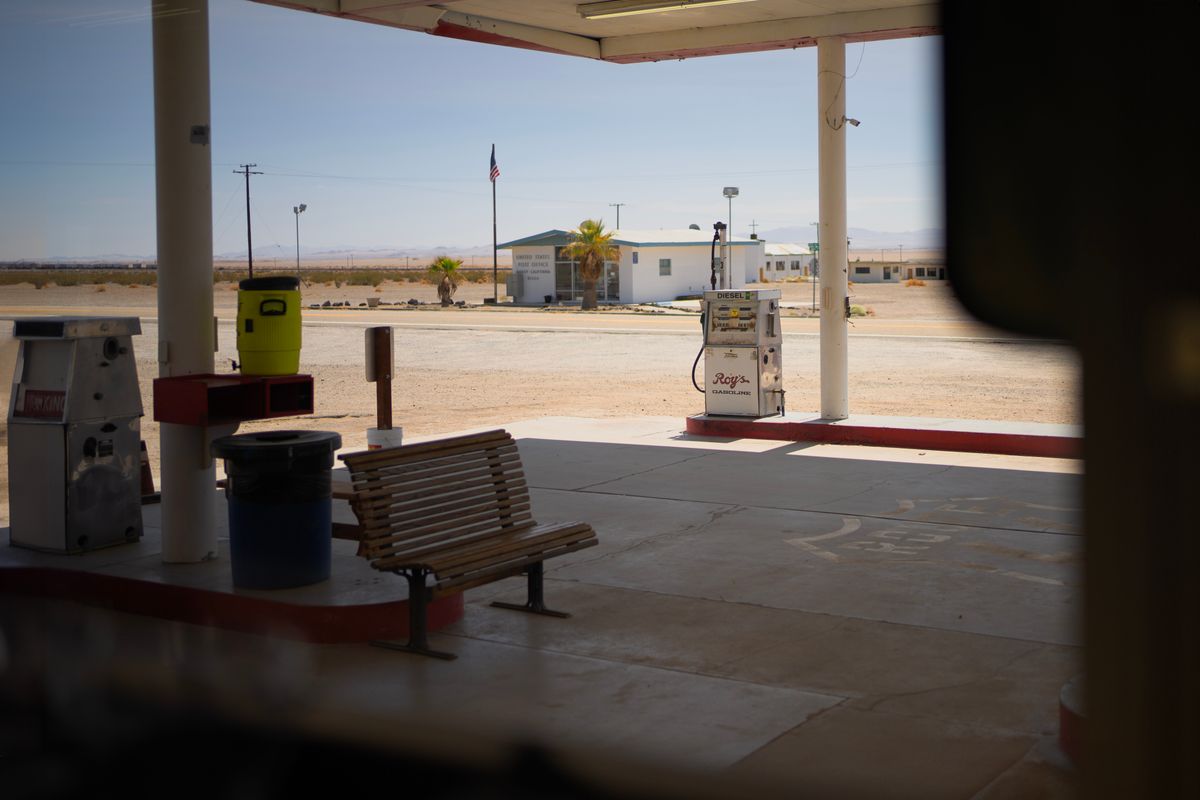 View from under the shaded canopy at Roy's Motel & Café in Amboy, a vintage gas pump and wooden bench in the foreground, a US flag, white desert outpost buildings, and Mojave flats stretching out behind.