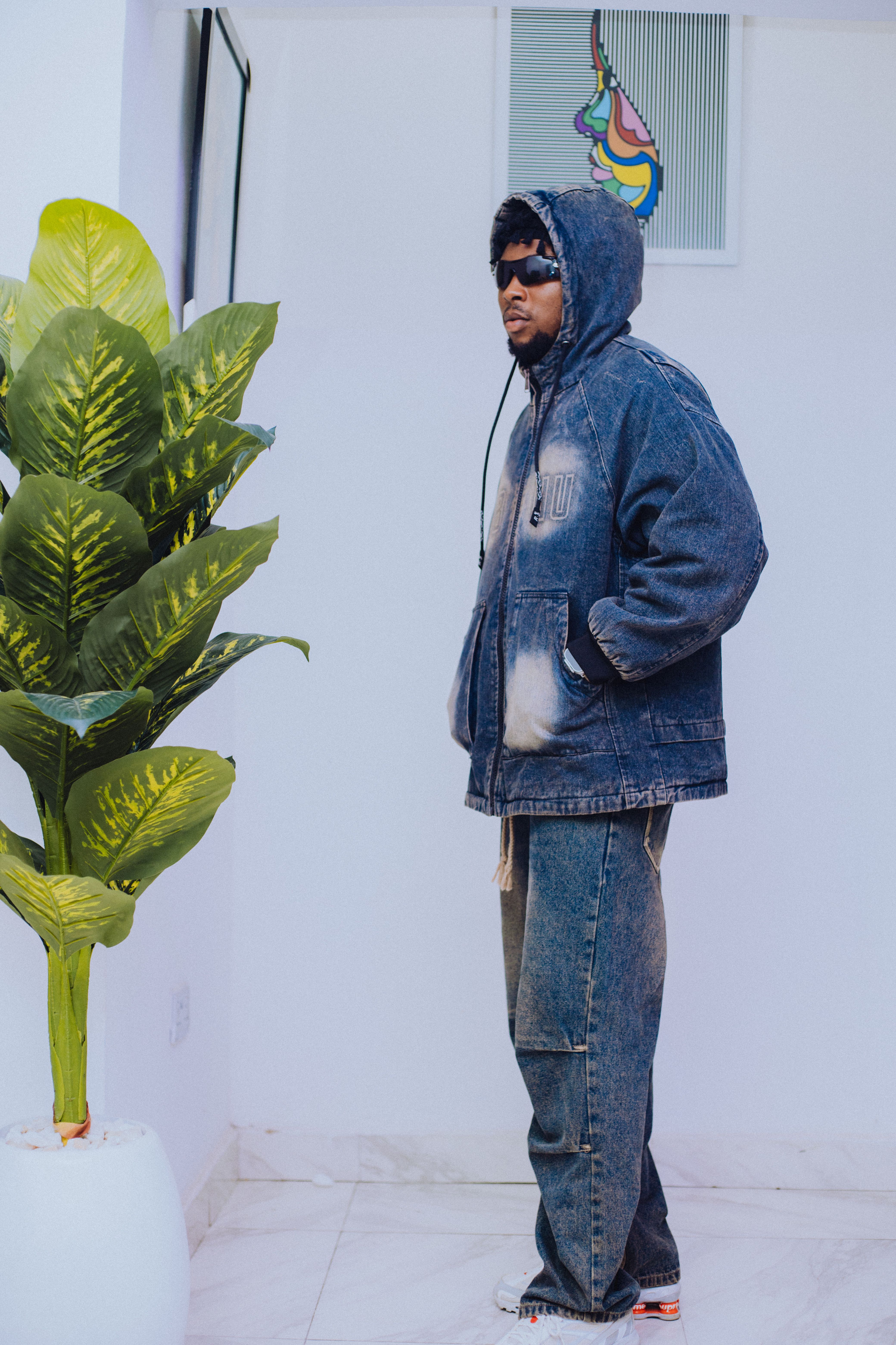 Man in denim outfit stands indoors with potted plant, sporting sunglasses.