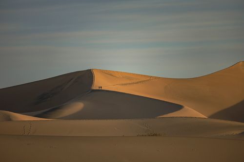 Free Silhouetted travelers walking along golden sand dunes under a vast sky at sunset. Stock Photo