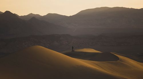 Free A lone figure stands atop sunlit sand dunes with mountains in the background at sunset. Stock Photo