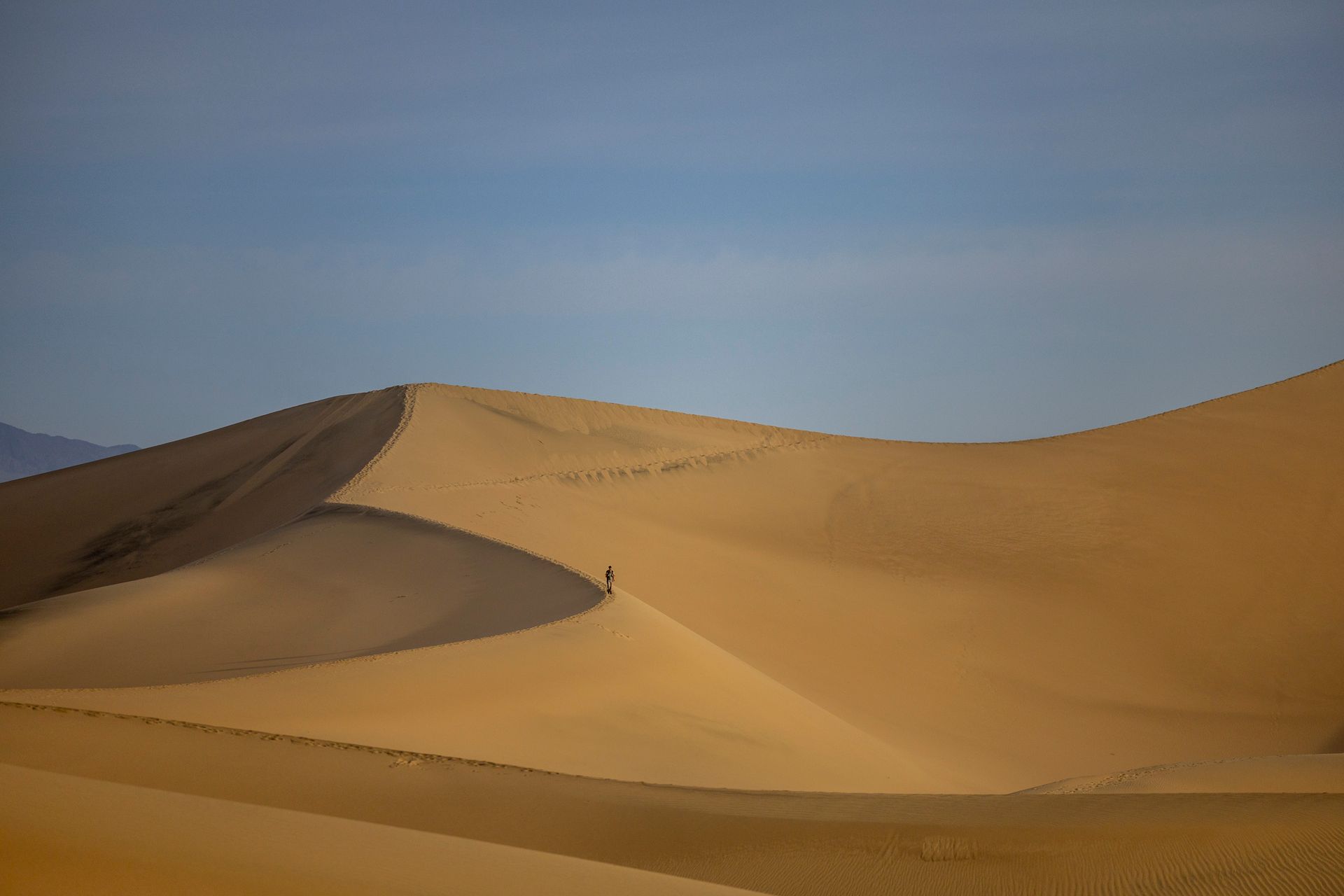 A lone figure traverses expansive desert sand dunes under a clear blue sky.