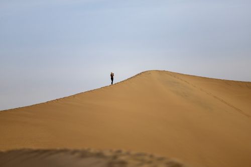 Free A solitary figure stands atop a vast desert dune under a clear blue sky. Stock Photo