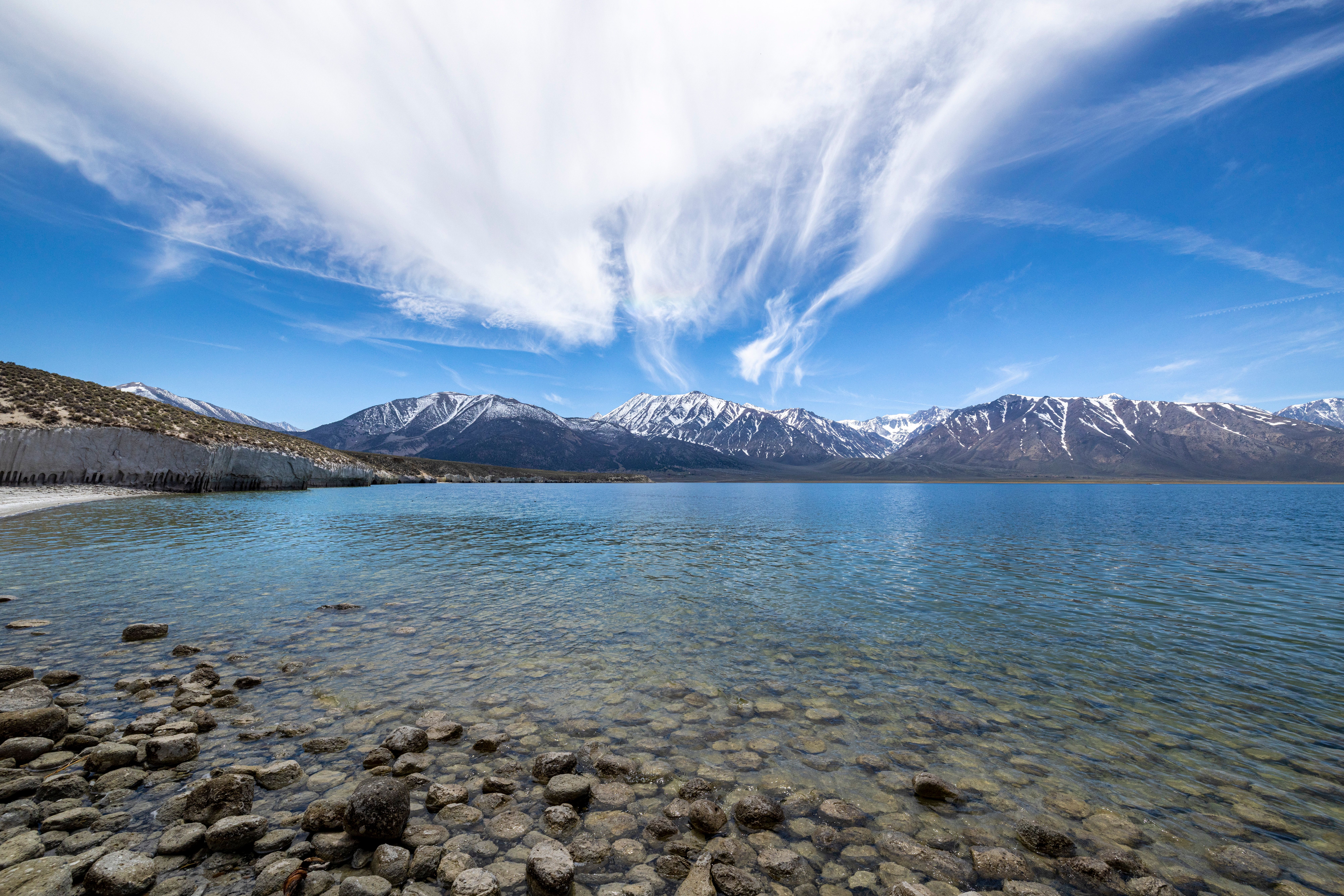 Stunning view of a tranquil lake with rocky foreground and snow-capped mountains under clear blue skies.