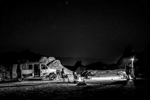 Black and white photo of desert camping under starry night sky with van and campfire.