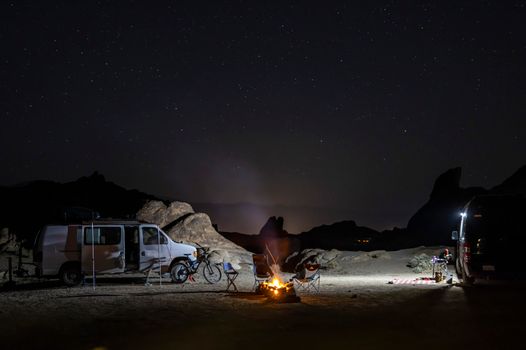 Serene night campsite with vans, chairs, and a campfire under a starry sky.