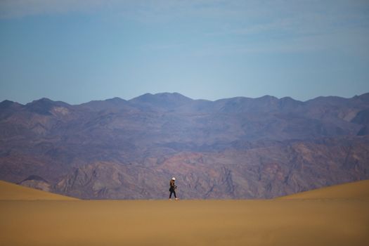 Person walking alone across sand dunes with a mountainous backdrop, showcasing solitude and exploration.