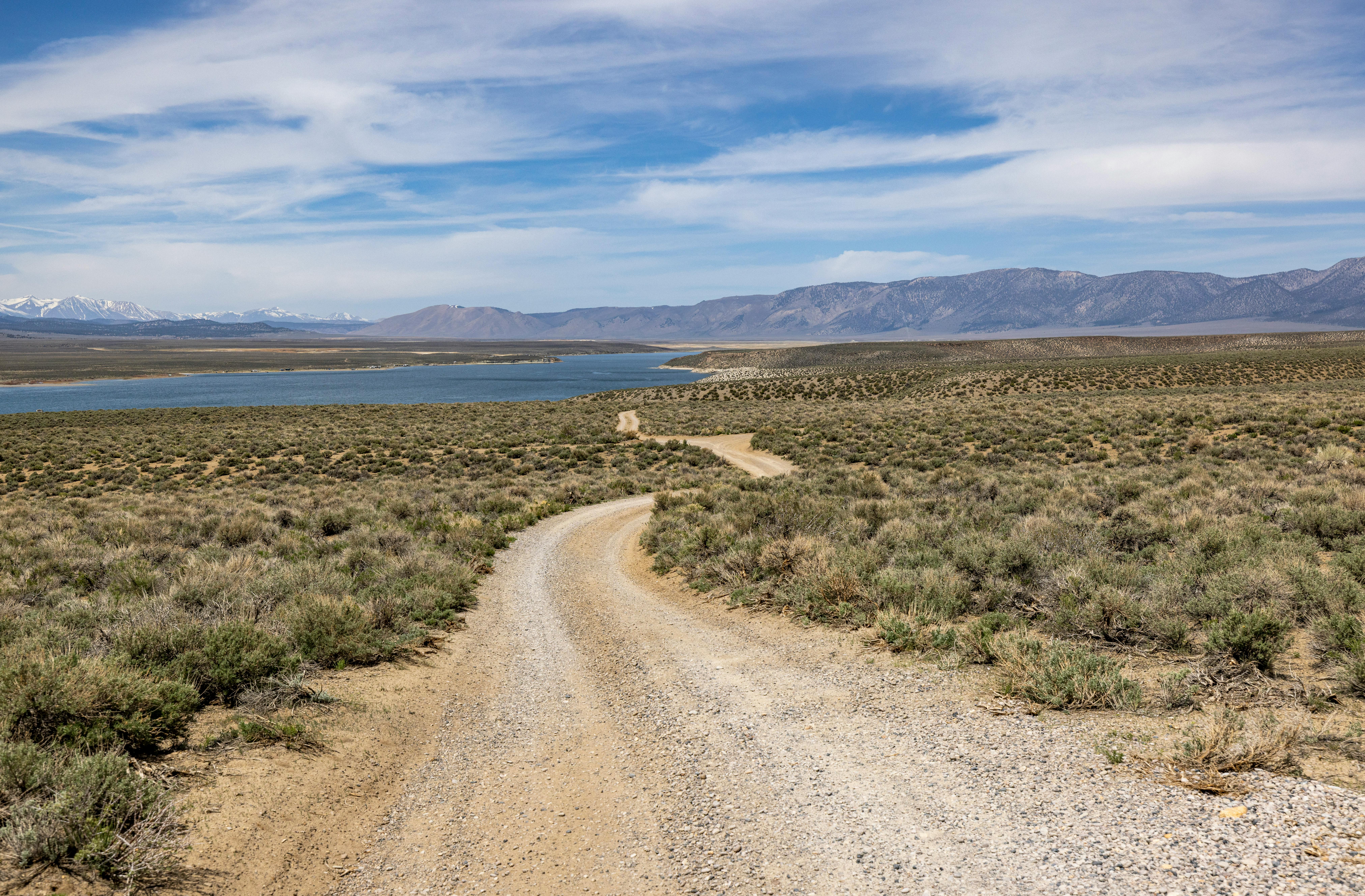 A winding dirt road leading to a tranquil lake surrounded by mountains under a vast blue sky.