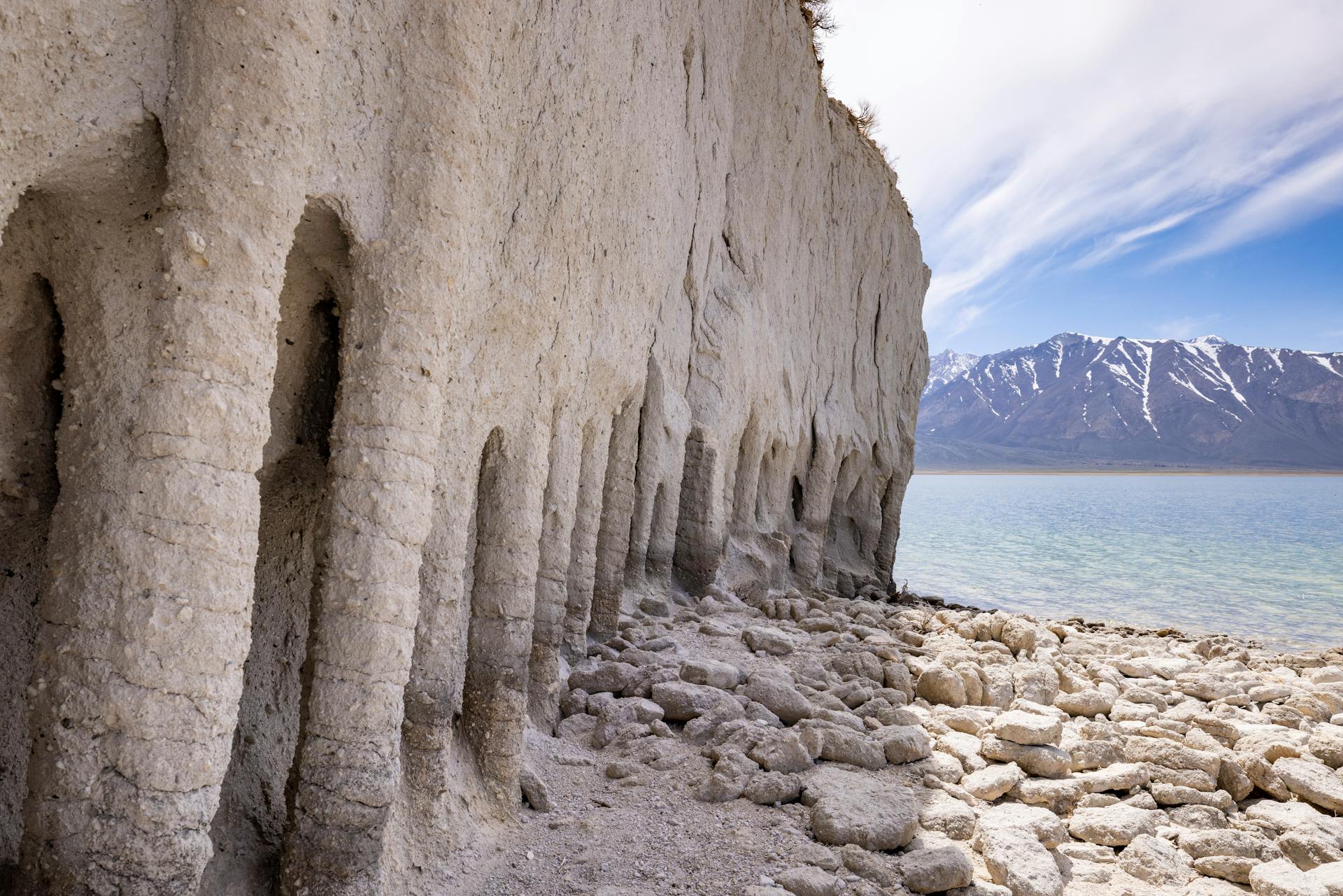 Eroded limestone formations by a tranquil lake with distant snow-capped mountains under a bright sky.